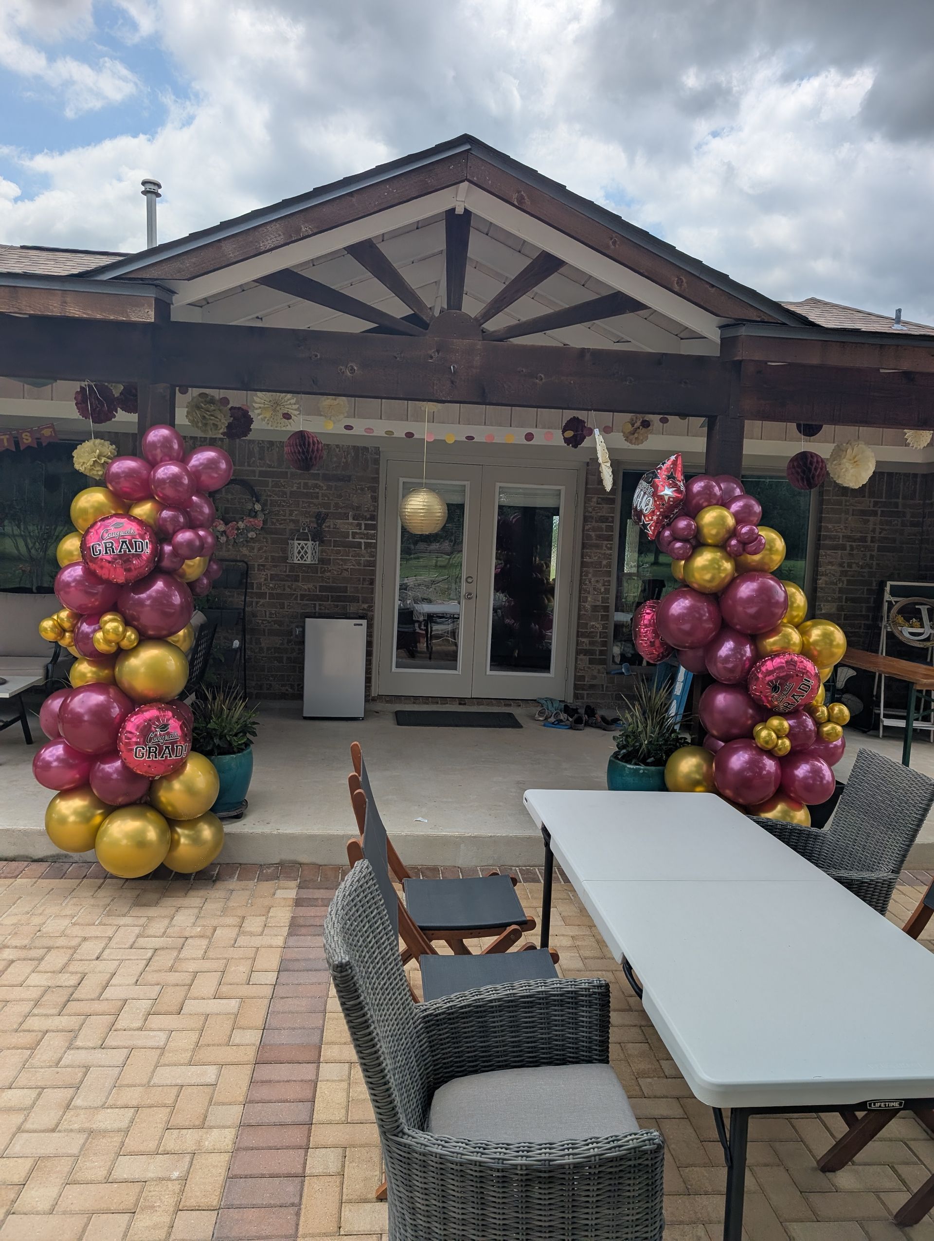 Party entrance with balloon columns in gold and burgundy, with a table and chairs on a patio.