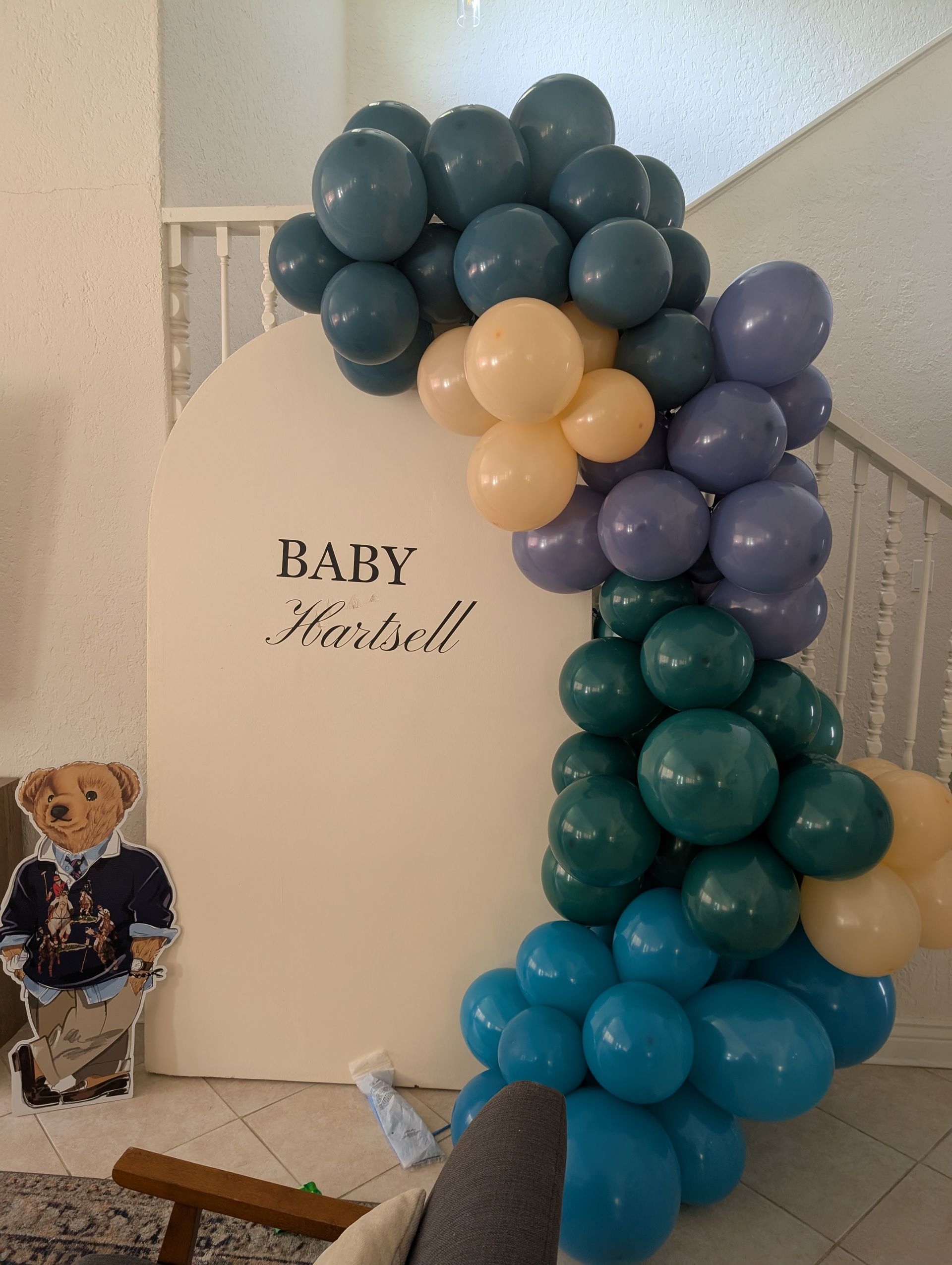 Baby shower backdrop with balloons in shades of blue, green, and beige. Teddy bear cutout present.