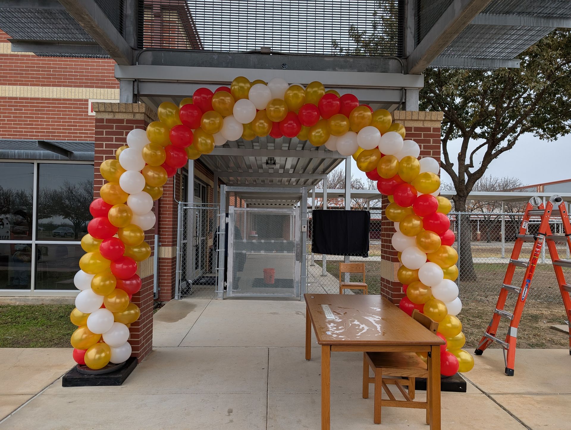 Balloon arch in red, yellow, and white, over a doorway with a table and ladder nearby.