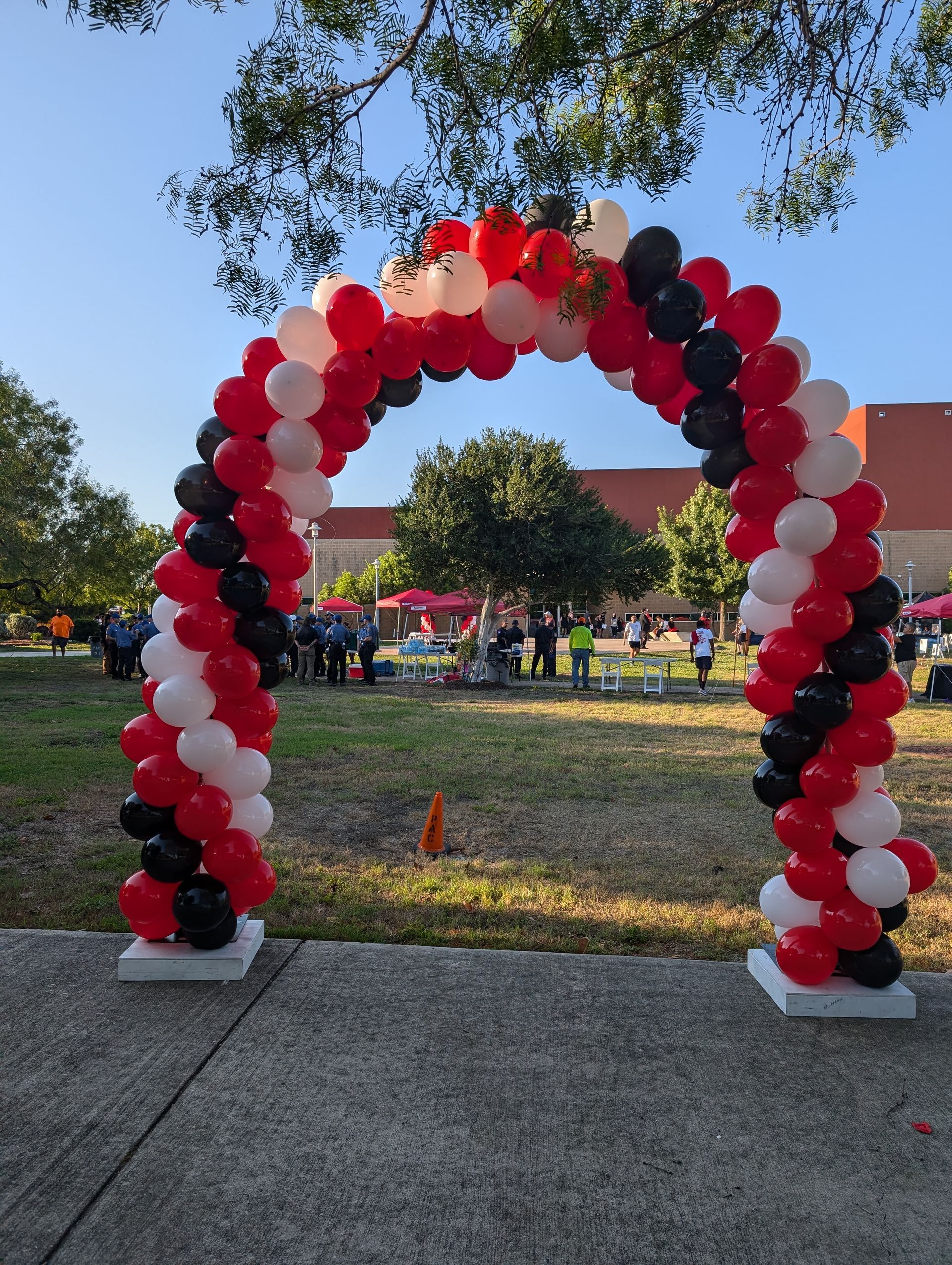 Balloon arch in red, white, and black colors, set up on a sidewalk with a park and people in the background.