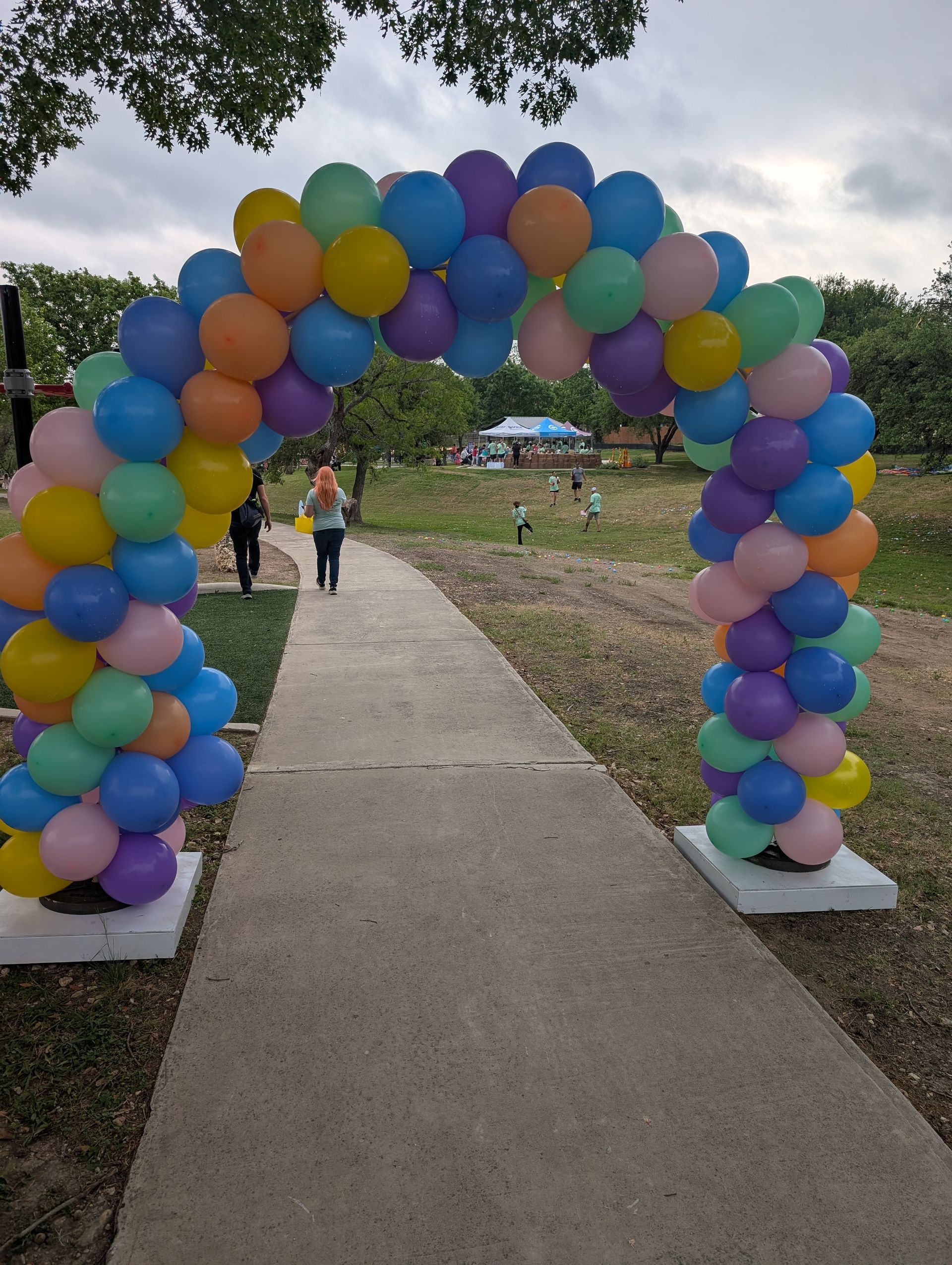 Archway of colorful balloons over a path, people walking towards a picnic area.