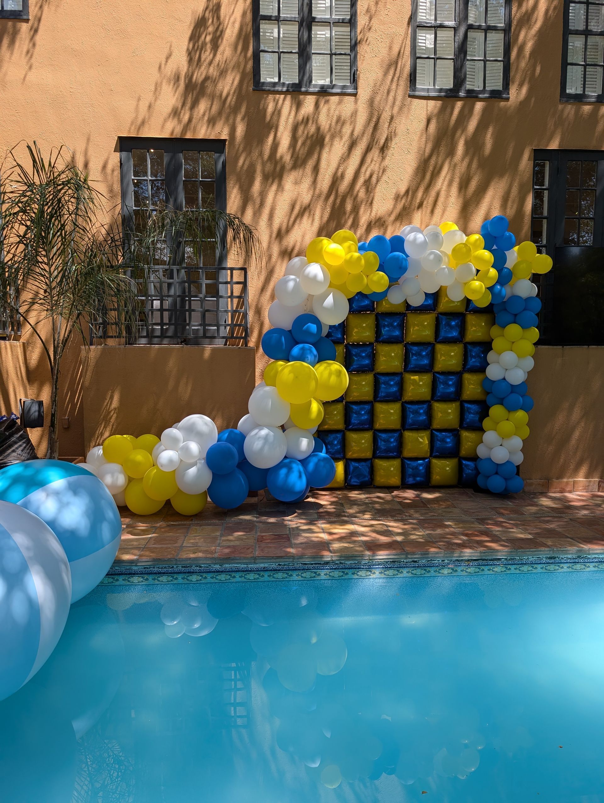 Poolside balloon arch in blue, yellow, and white with checkerboard design; sunny day.
