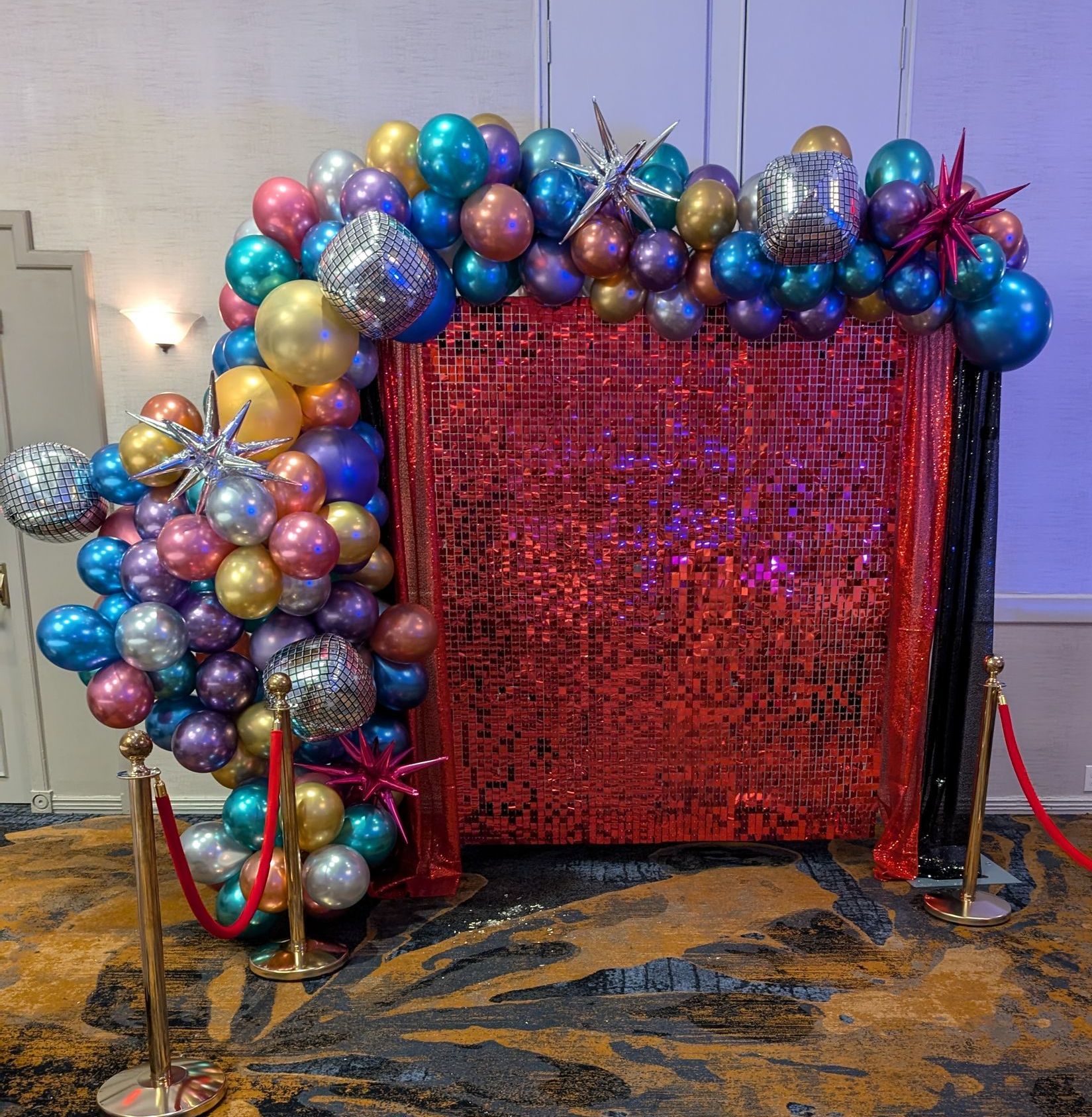 A photo booth with a red sequined backdrop and a colorful balloon archway, indoors.
