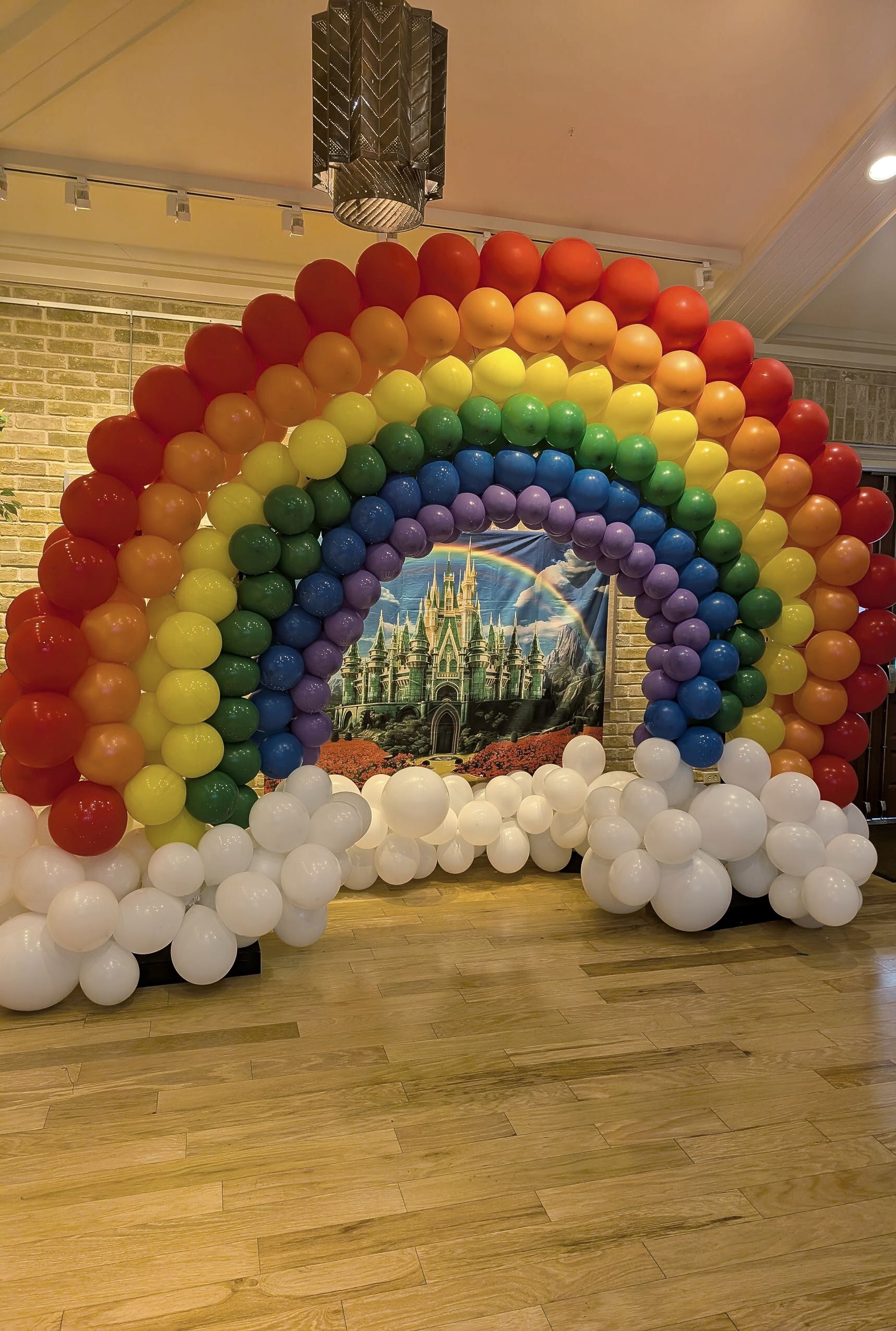 Rainbow-shaped balloon arch with white clouds at the base, framing a castle backdrop.