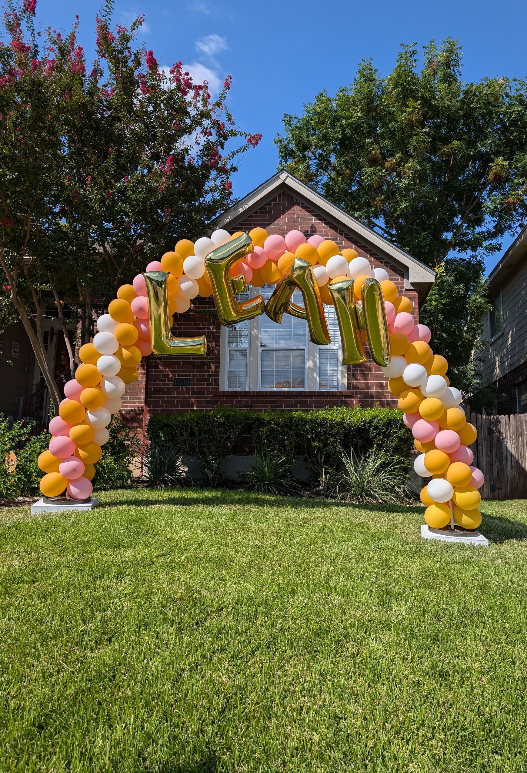 Balloon arch with "LEAH" in gold against a house. Yellow, white, and peach balloons arch over green grass.