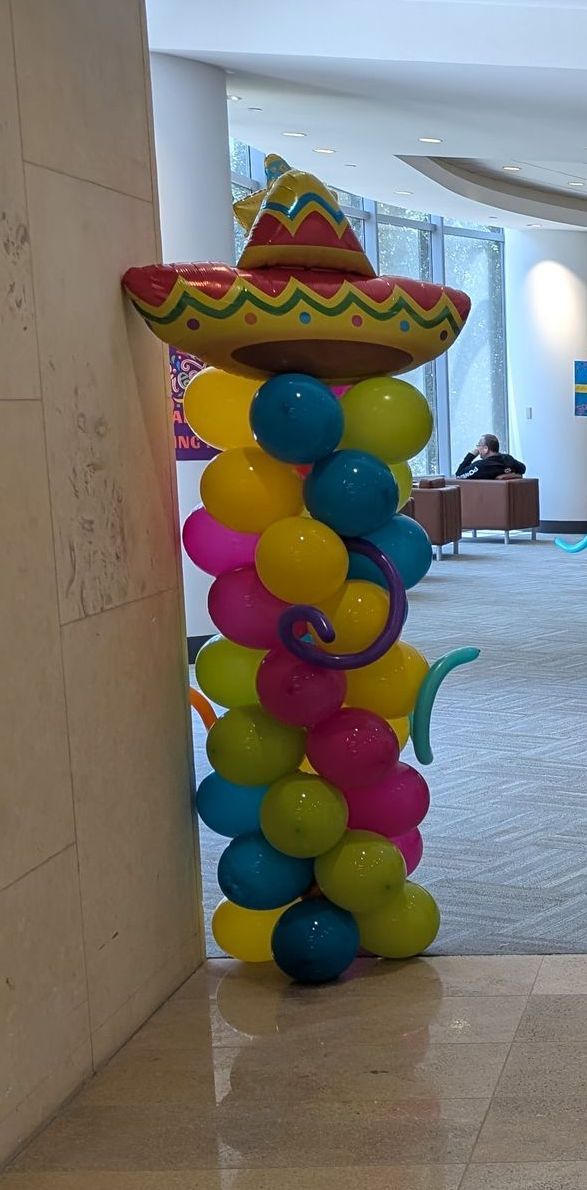 Balloon column decoration with a sombrero. Colors: yellow, blue, pink, green, and red. Placed by a wall.