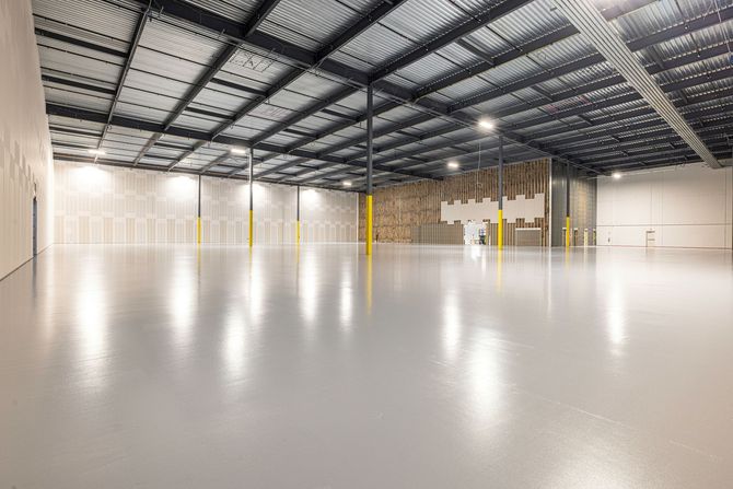 A vast, empty warehouse with a high, dark steel ceiling, yellow support pillars, and a polished, light-gray epoxy floor.