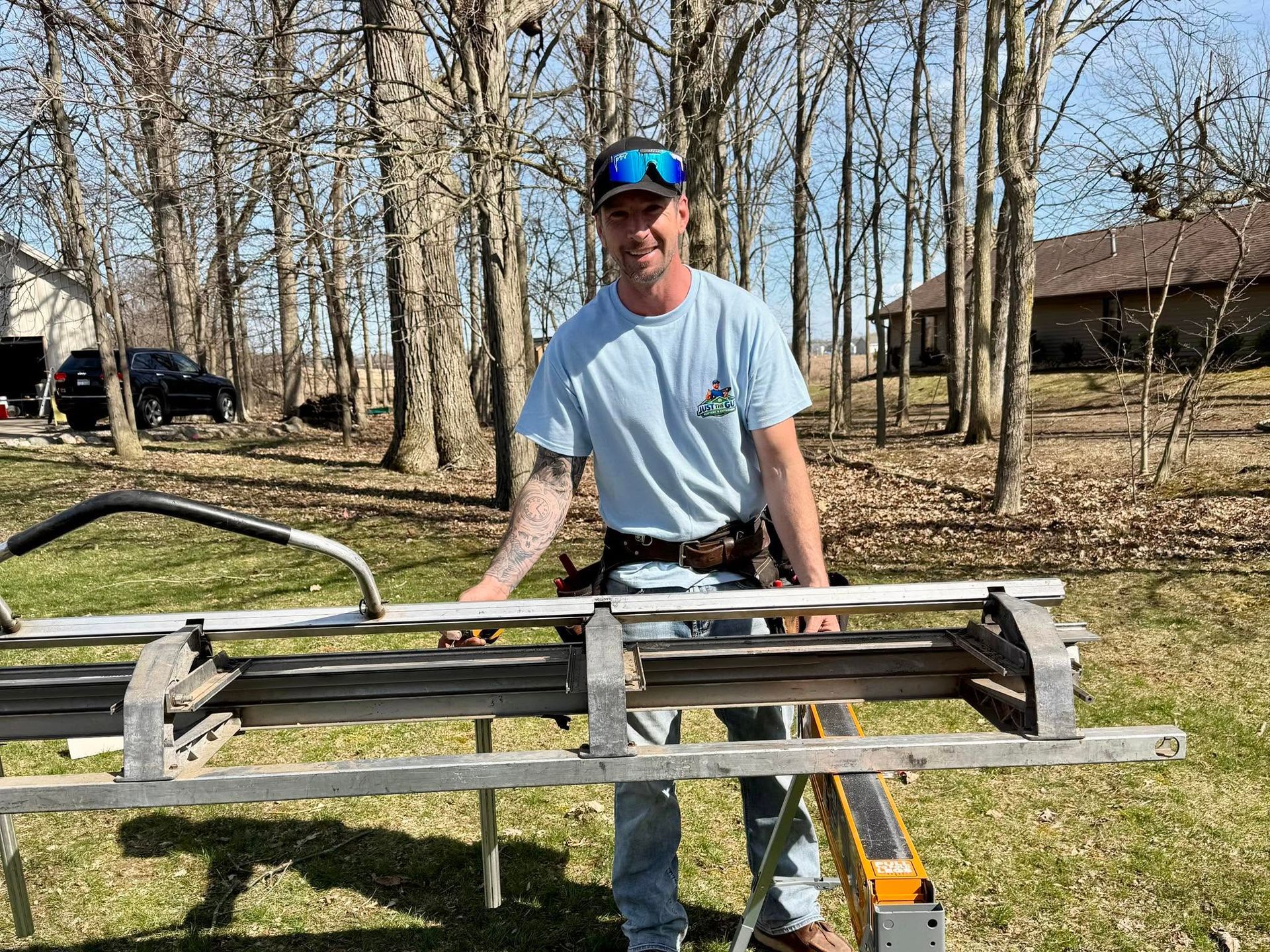 Man standing outdoors beside a wooden frame on a saw, holding tools in a yard with leafless trees.