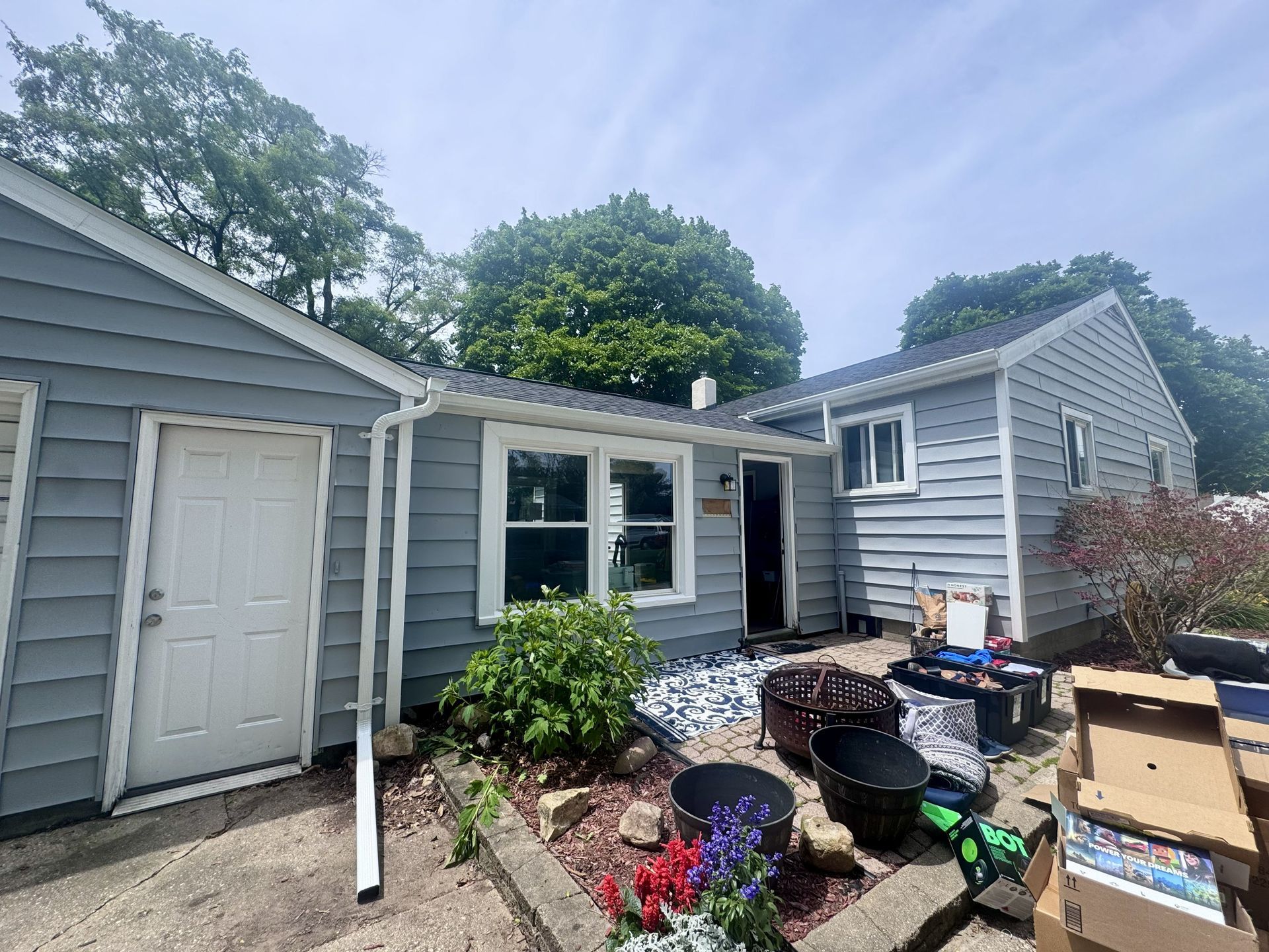 Backyard patio beside gray house, with open doorway, potted plants, and boxes scattered on the ground