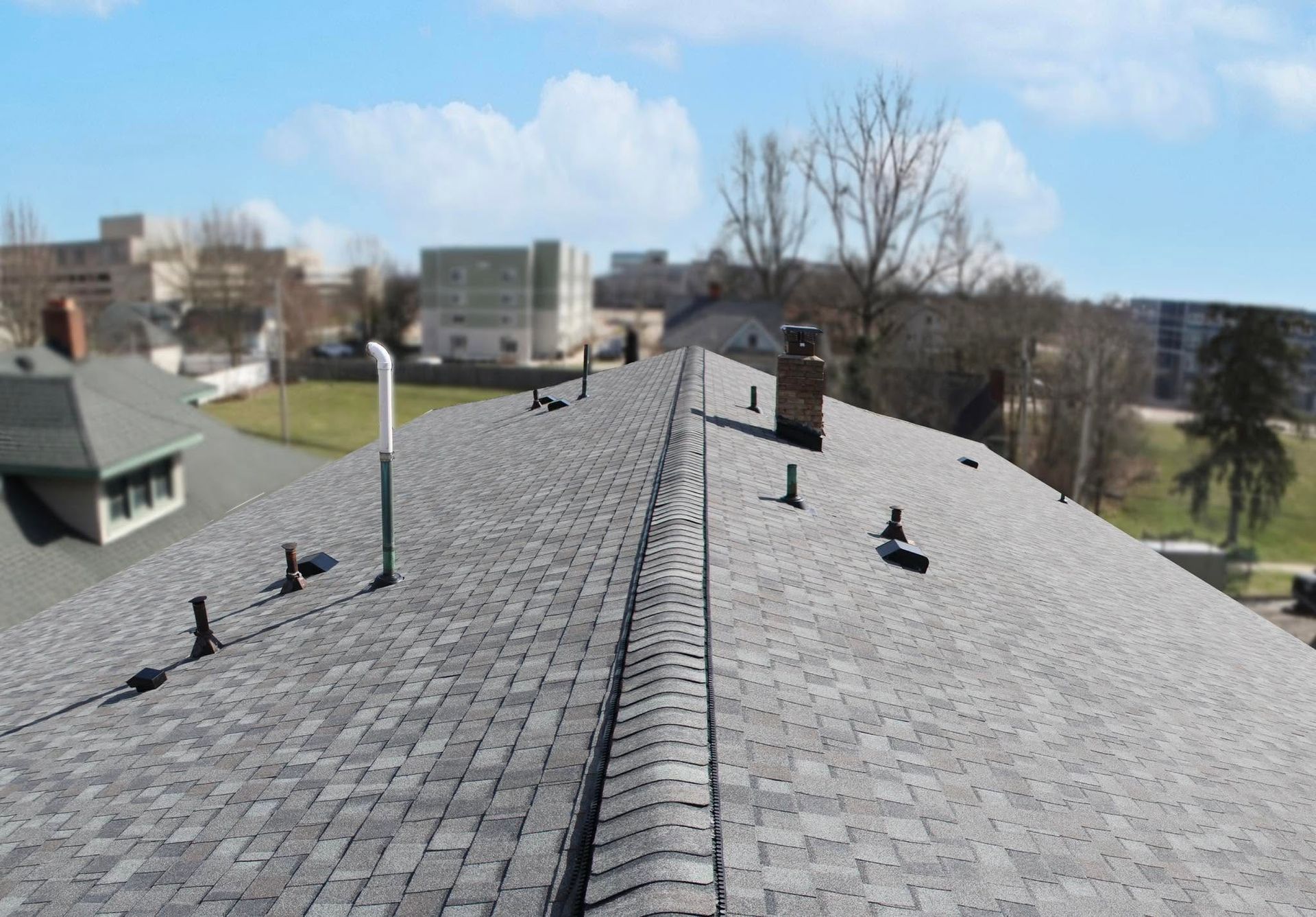 Rooftop with dark gray shingles and vents, overlooking buildings and trees on a clear day