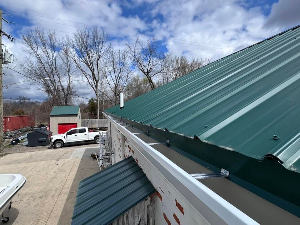 Green metal roof on a building beside a yard with parked vehicles and leafless trees under a cloudy sky
