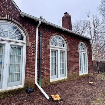 Brick house exterior with three arched windows, white trim, and a white downspout in a muddy yard.