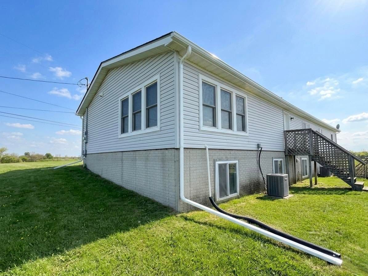 White house with vinyl siding on a grassy hill, showing side windows, basement, and exterior stairs under a blue sky