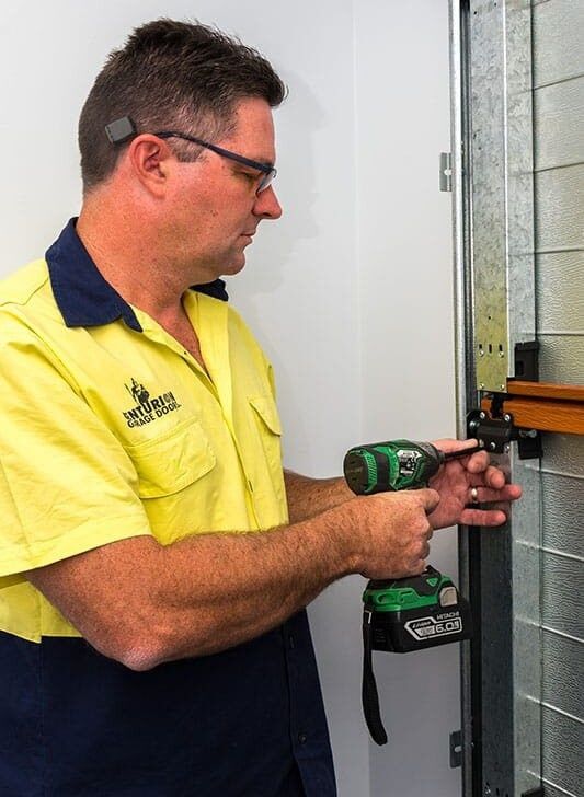 A Man in A Yellow Shirt Is Working on A Door with A Drill — Gem Garage Doors in Coffs Harbour, NSW