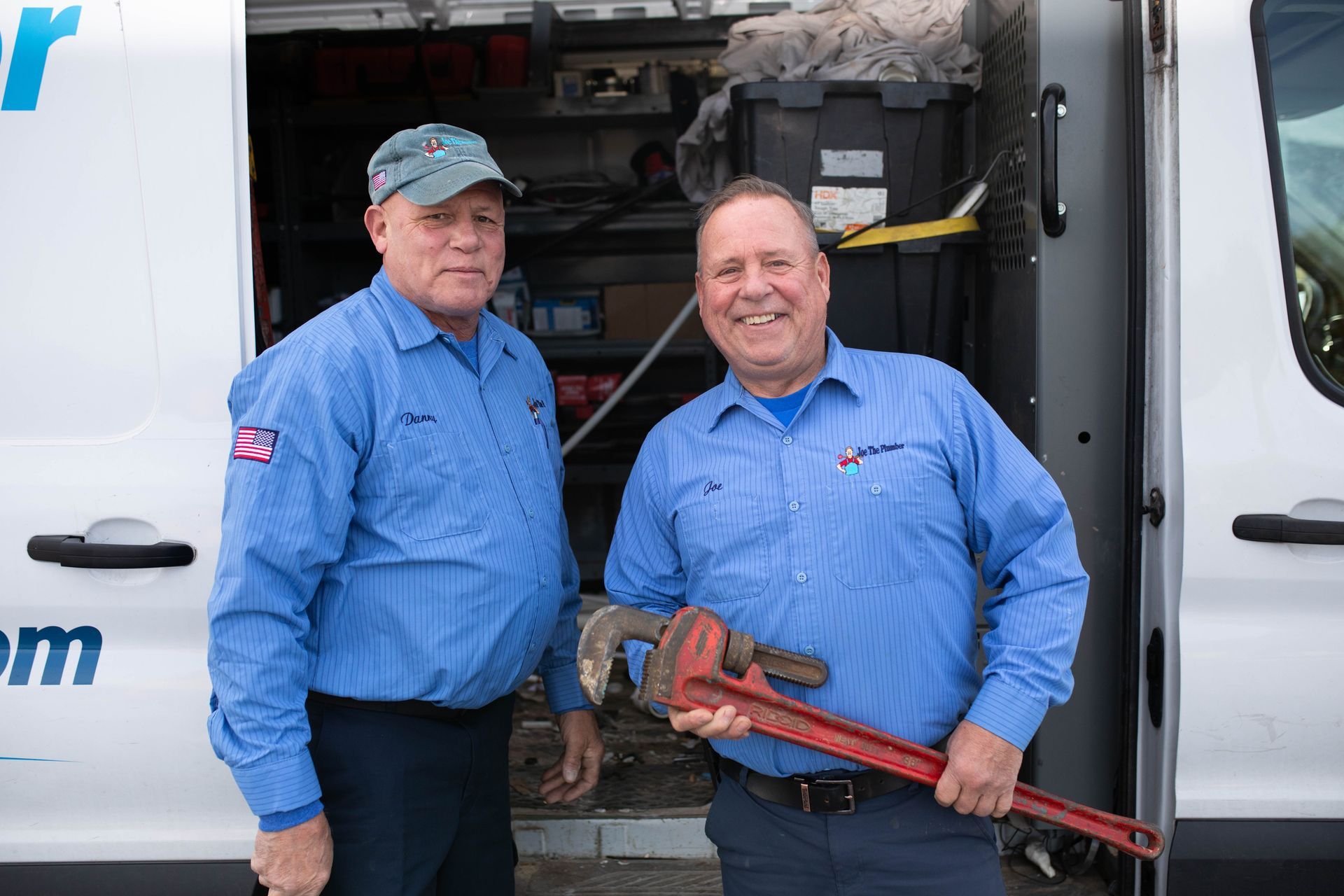 Two plumbers stand by a service van. One holds a large pipe wrench. Both wear blue uniforms and smiles.