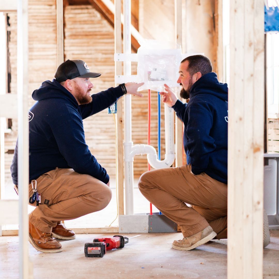 Two construction workers in dark hoodies and tan pants crouch in a wooden frame house, discussing plumbing installations.