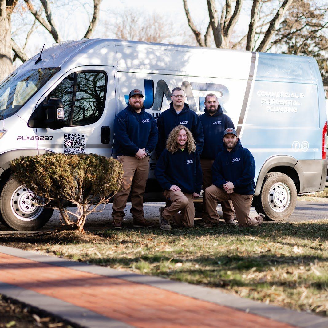 A group of five people in navy blue sweatshirts stand in front of a white JMR construction van on a sunny day.