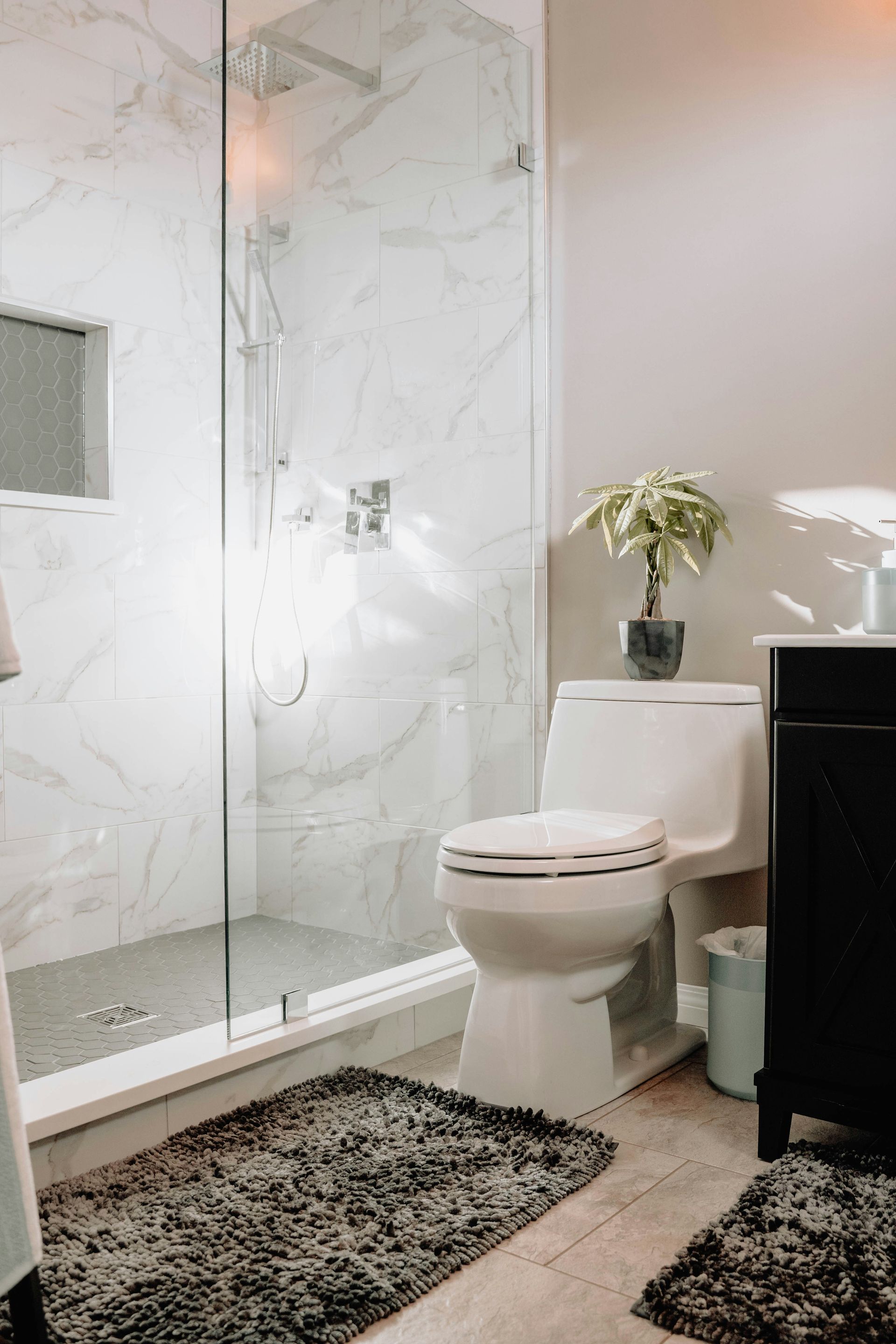 A modern bathroom featuring a white toilet, marble-tiled shower, dark vanity, and two dark gray shaggy floor mats.