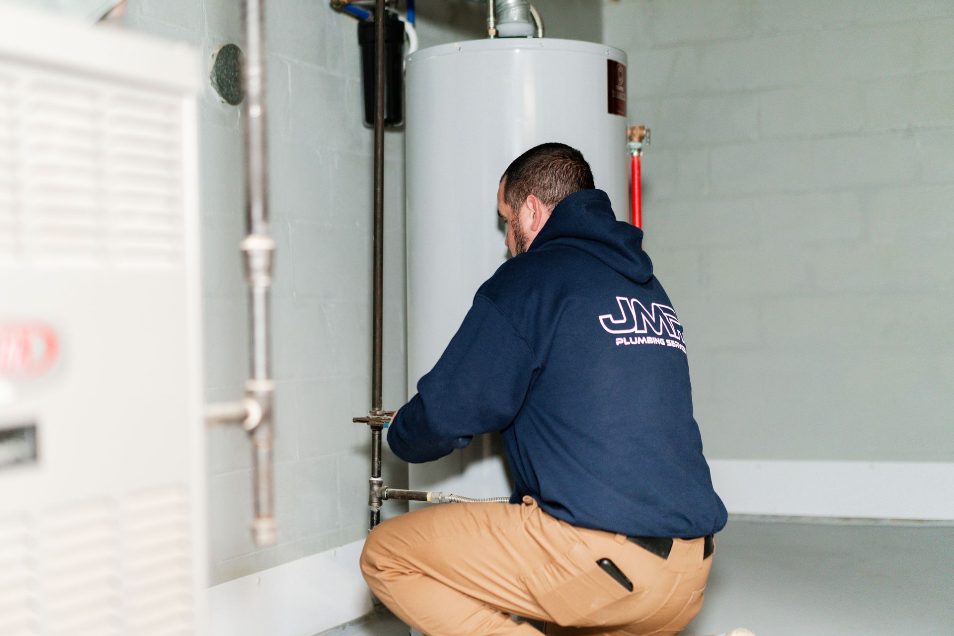A plumber wearing a dark blue hoodie and tan work pants kneels to repair a white water heater in a basement.