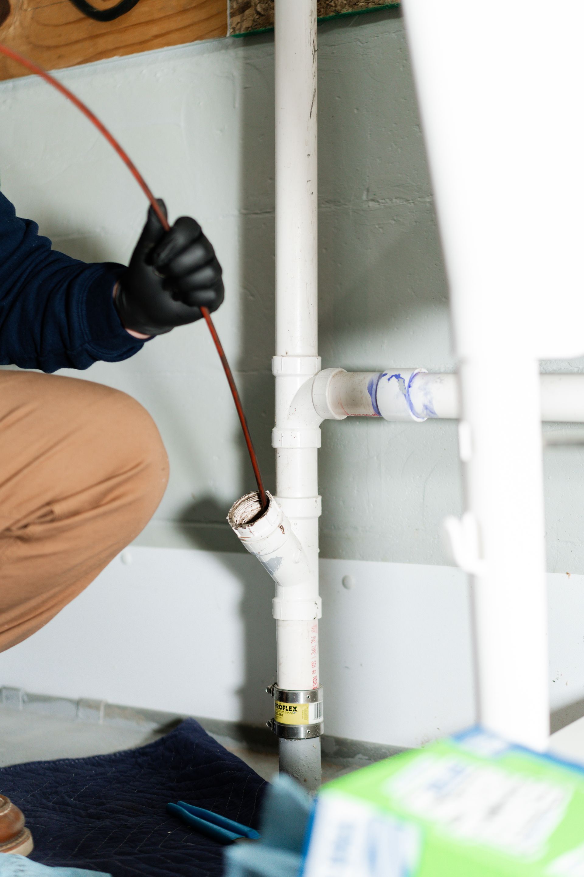 A plumber in black gloves inserts a drain snake into a white PVC pipe fitting against a basement wall.