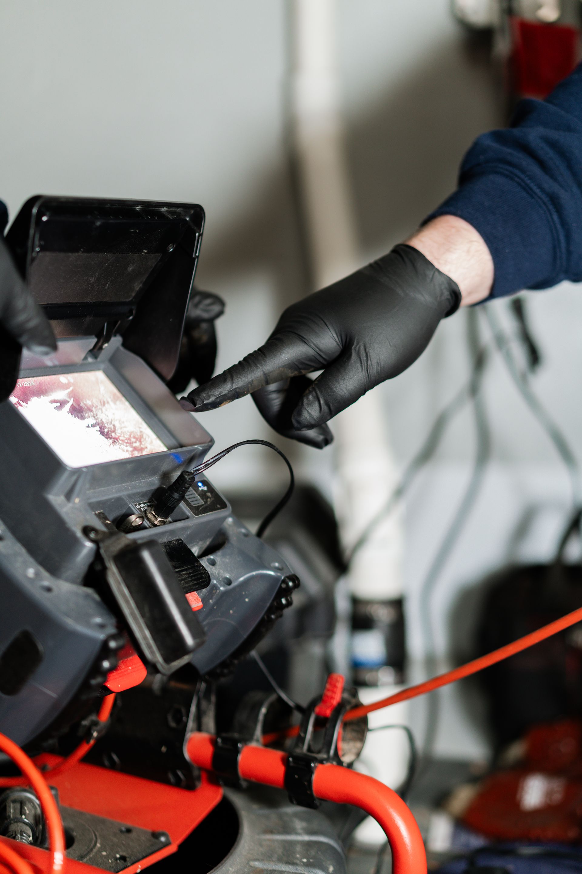 A person in black gloves points to a screen on a drain inspection camera display showing inside a pipe.