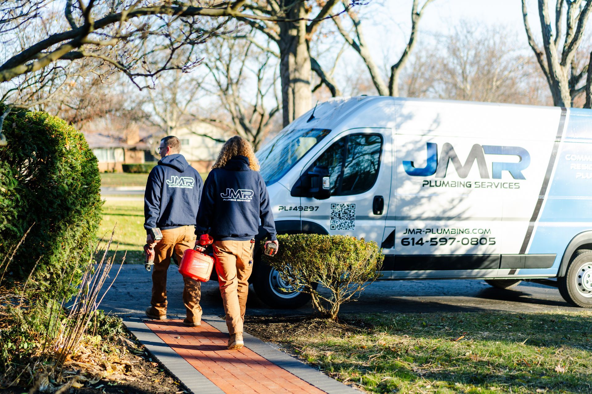 Two workers in dark blue branded jackets and tan pants walk toward a parked JMR Plumbing services van on a sunny day.