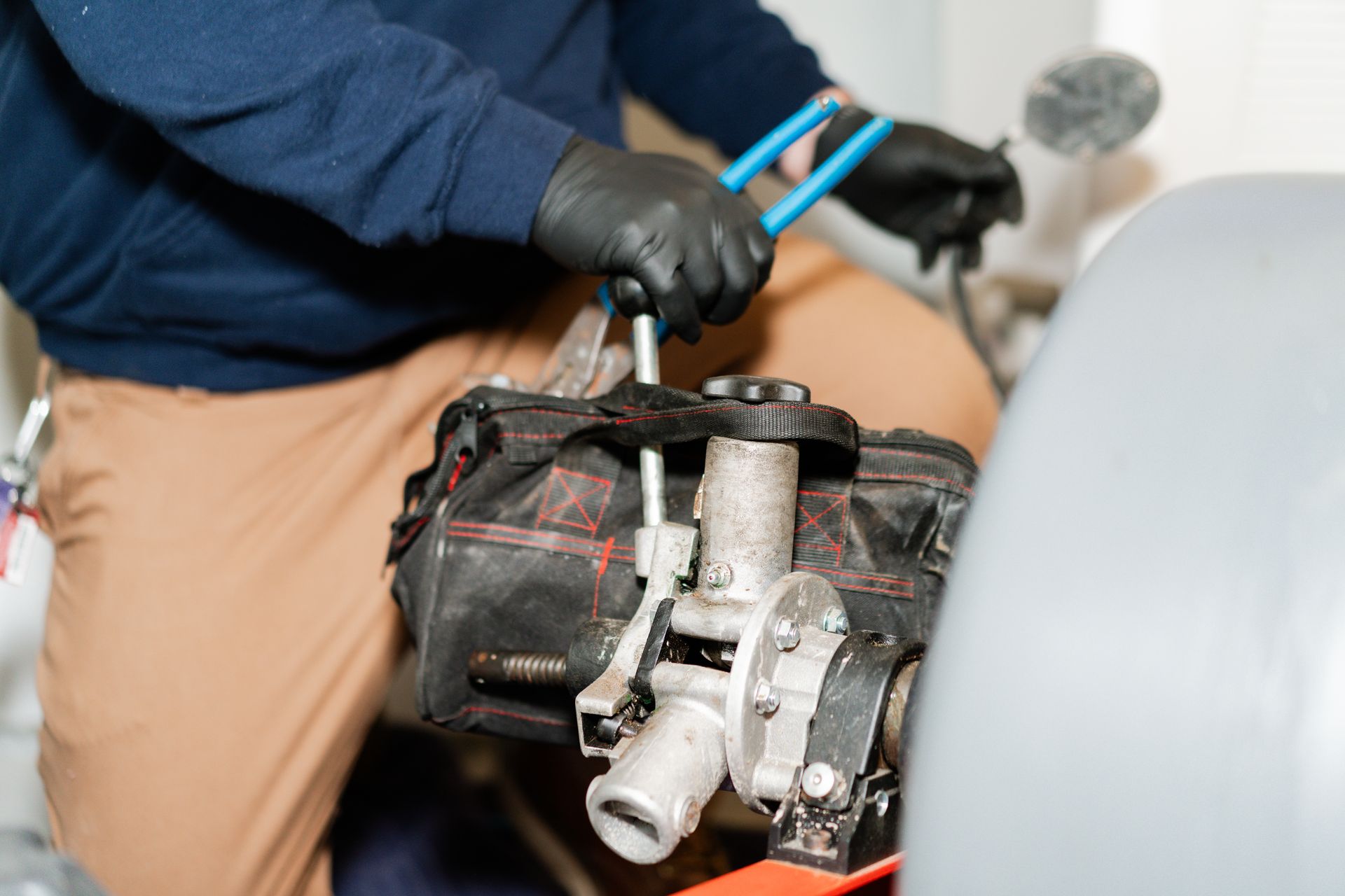 A person in a blue hoodie and black gloves uses blue-handled pliers to work on a mechanical part attached to a black bag.