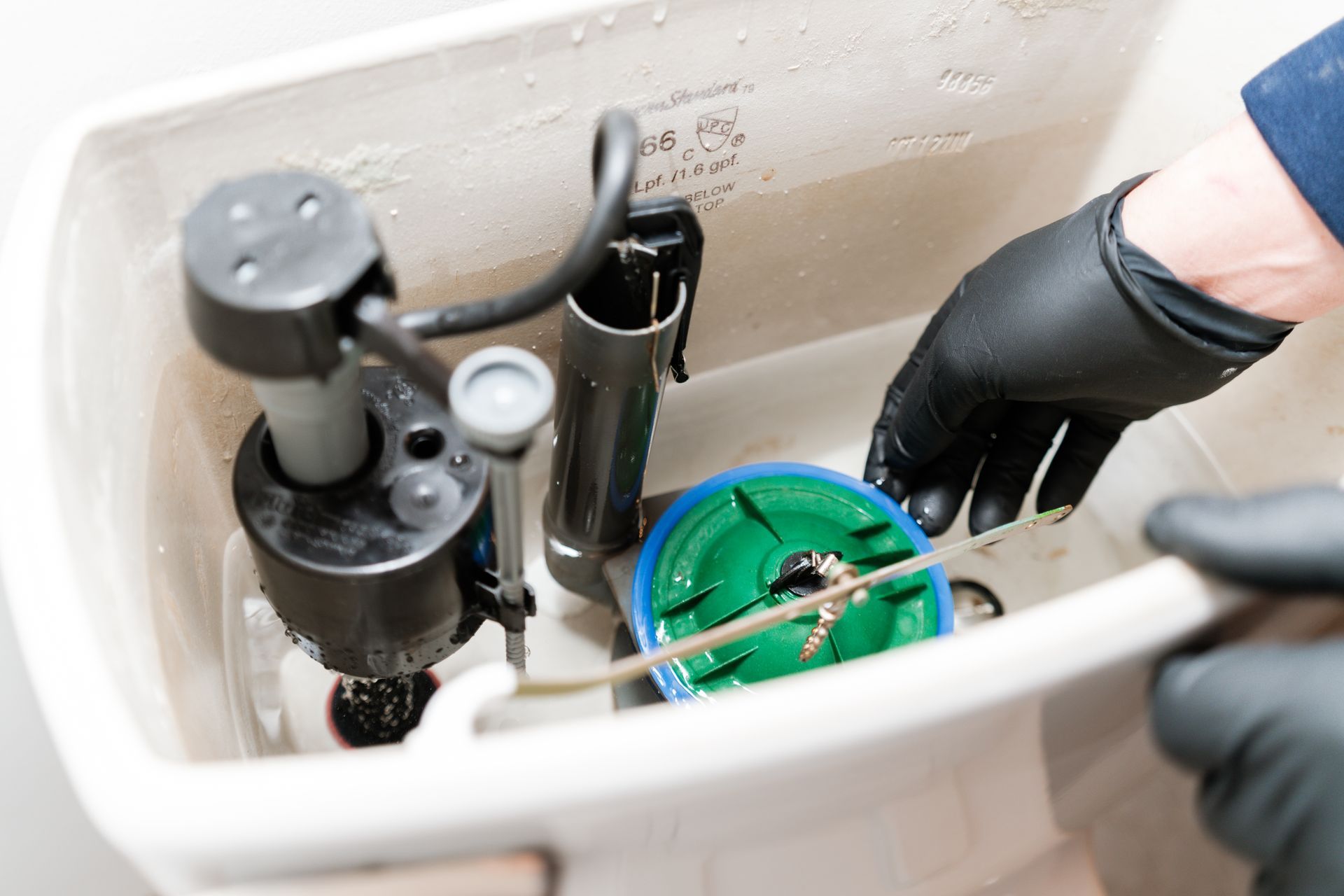 A person in black gloves reaches into an open toilet tank to adjust the green flush valve and chain.