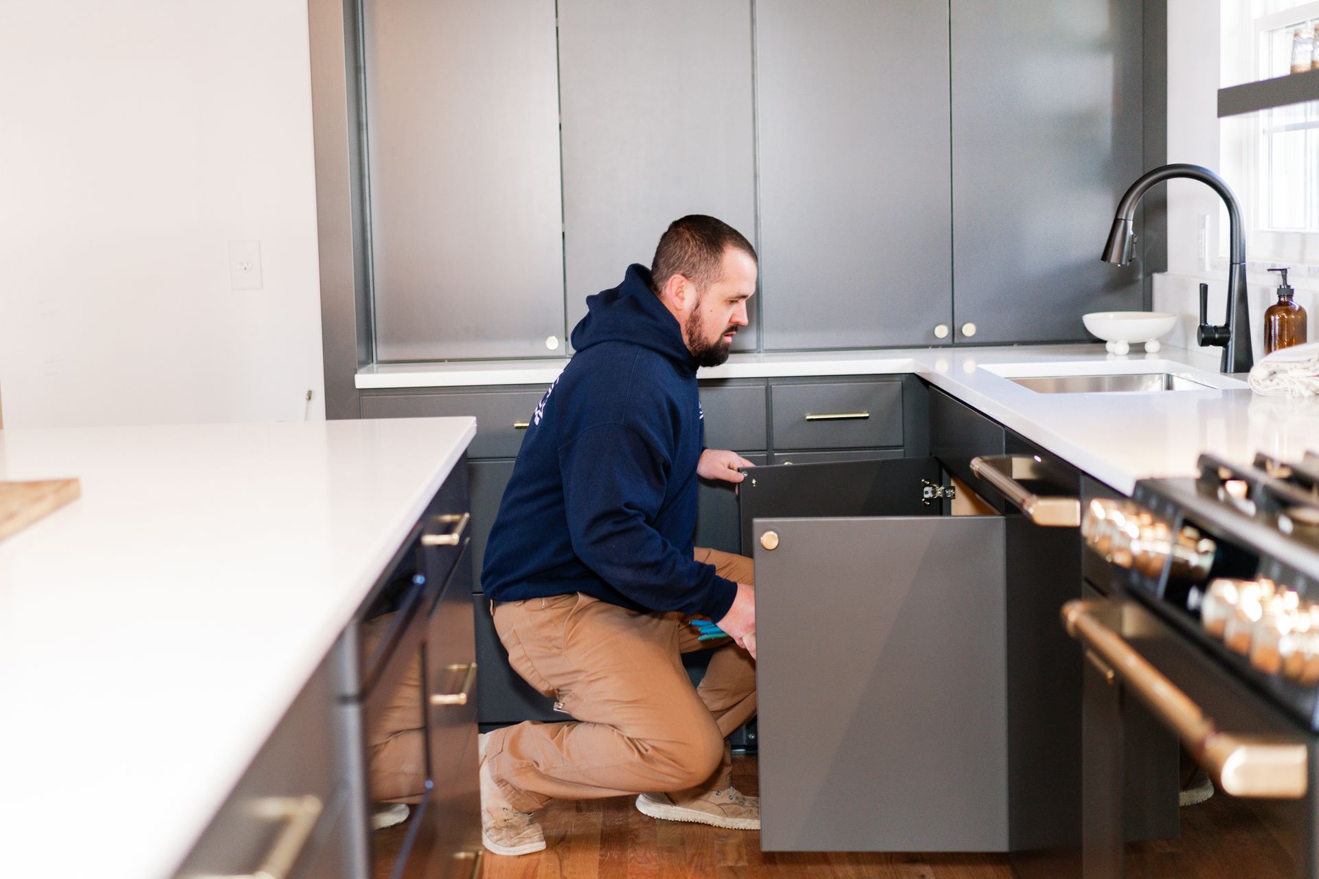 A person in a blue hoodie and tan pants crouches to inspect or work on the open lower cabinet of a grey kitchen island.