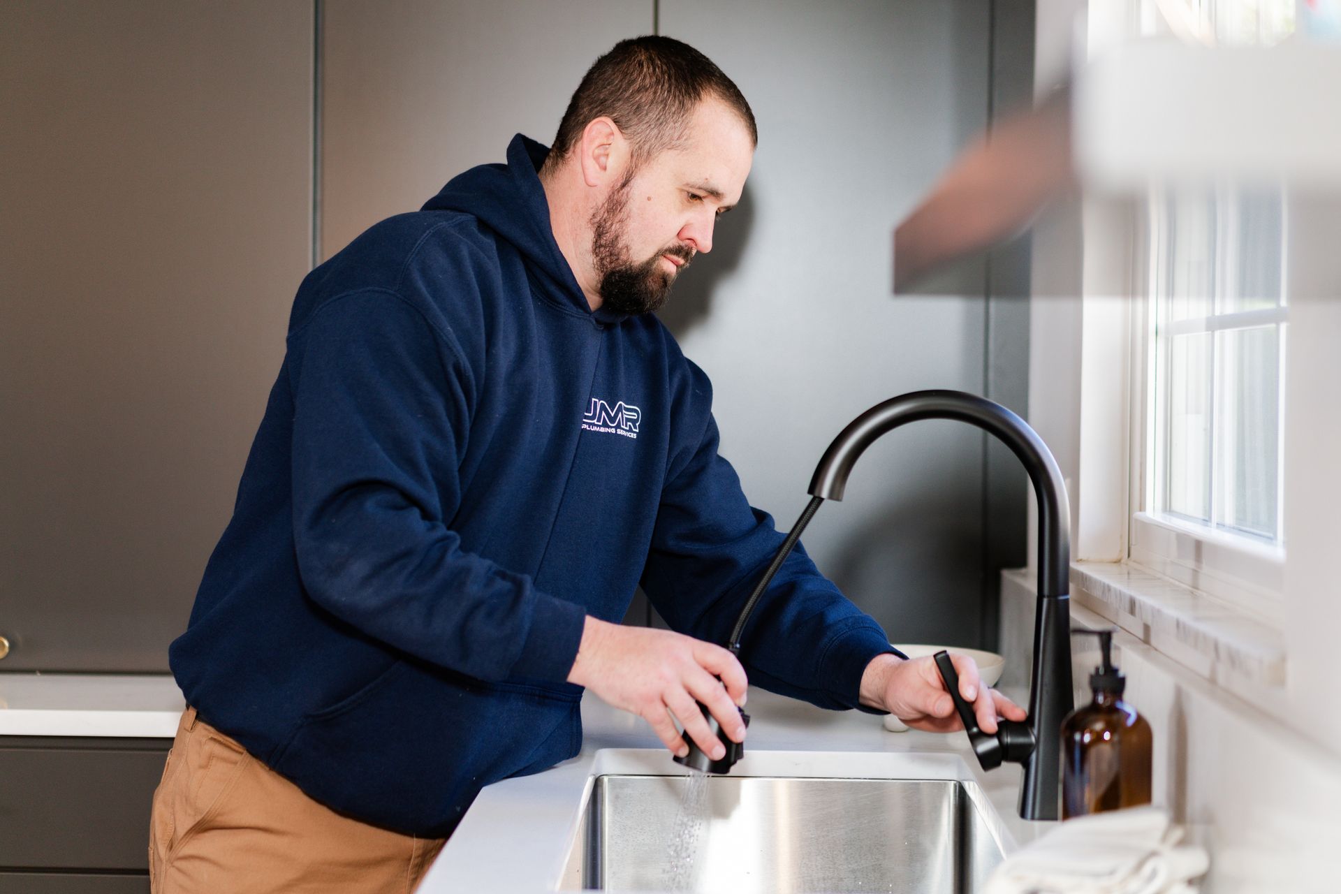 A professional in a navy hoodie repairs a modern kitchen faucet over a stainless steel sink.