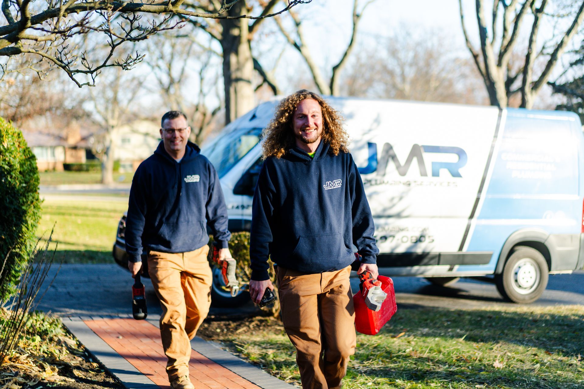 Two technicians in matching navy hoodies and tan pants walk toward the camera with tools, a company van parked behind them.