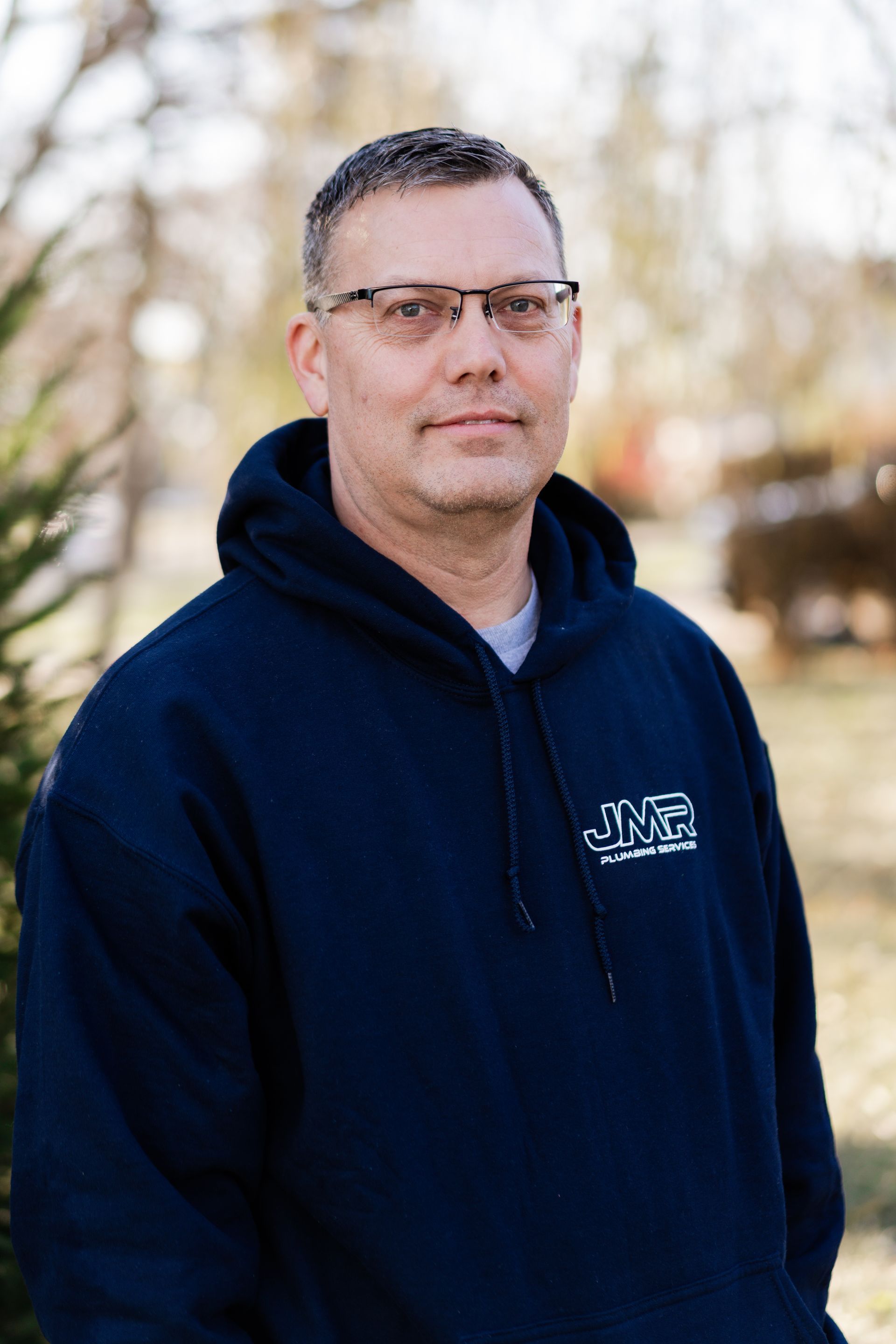 A person with short hair and glasses wearing a navy blue hoodie, standing outdoors in a soft-focus setting.