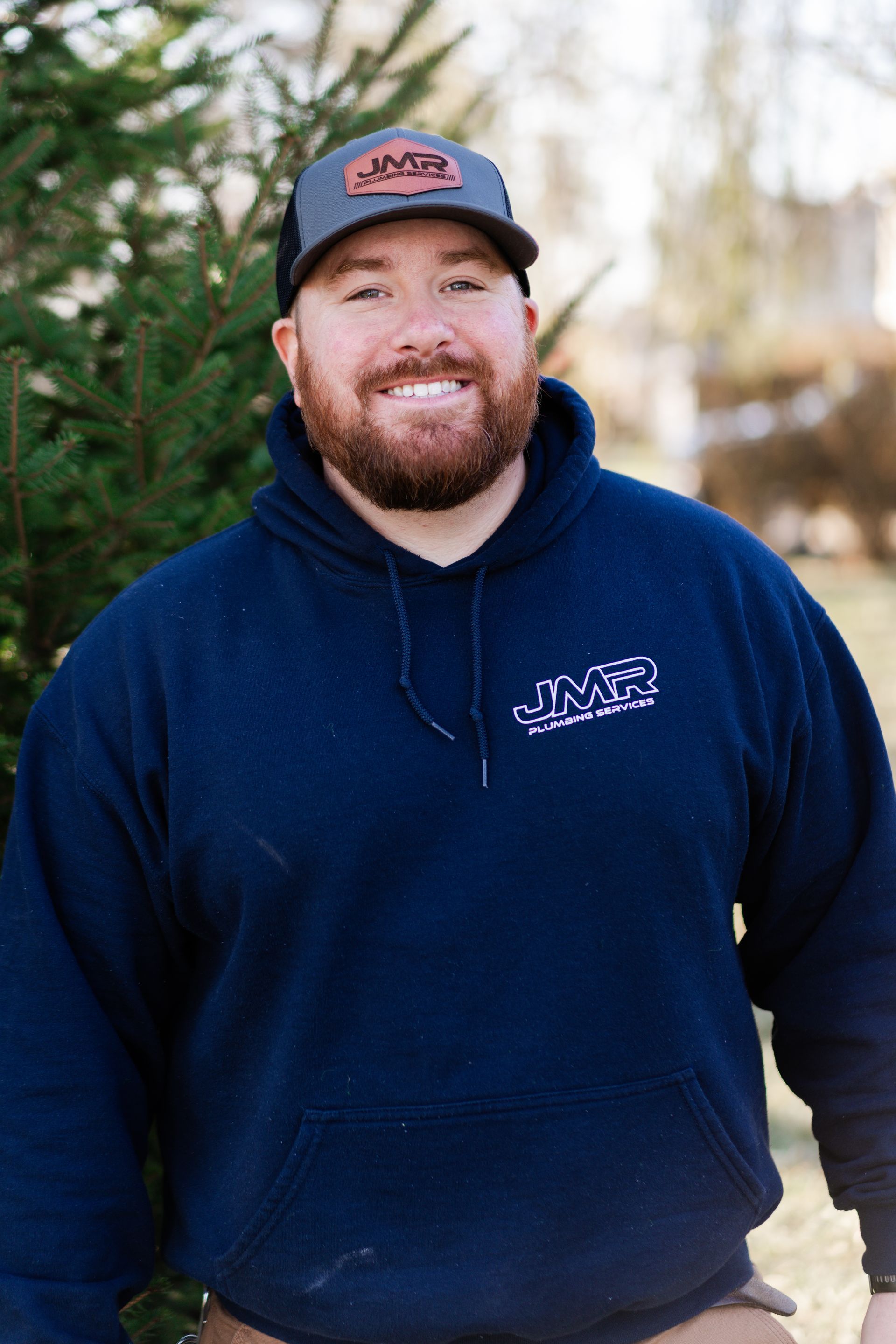 A smiling person in a blue hoodie and gray baseball cap standing outdoors near a conifer tree.