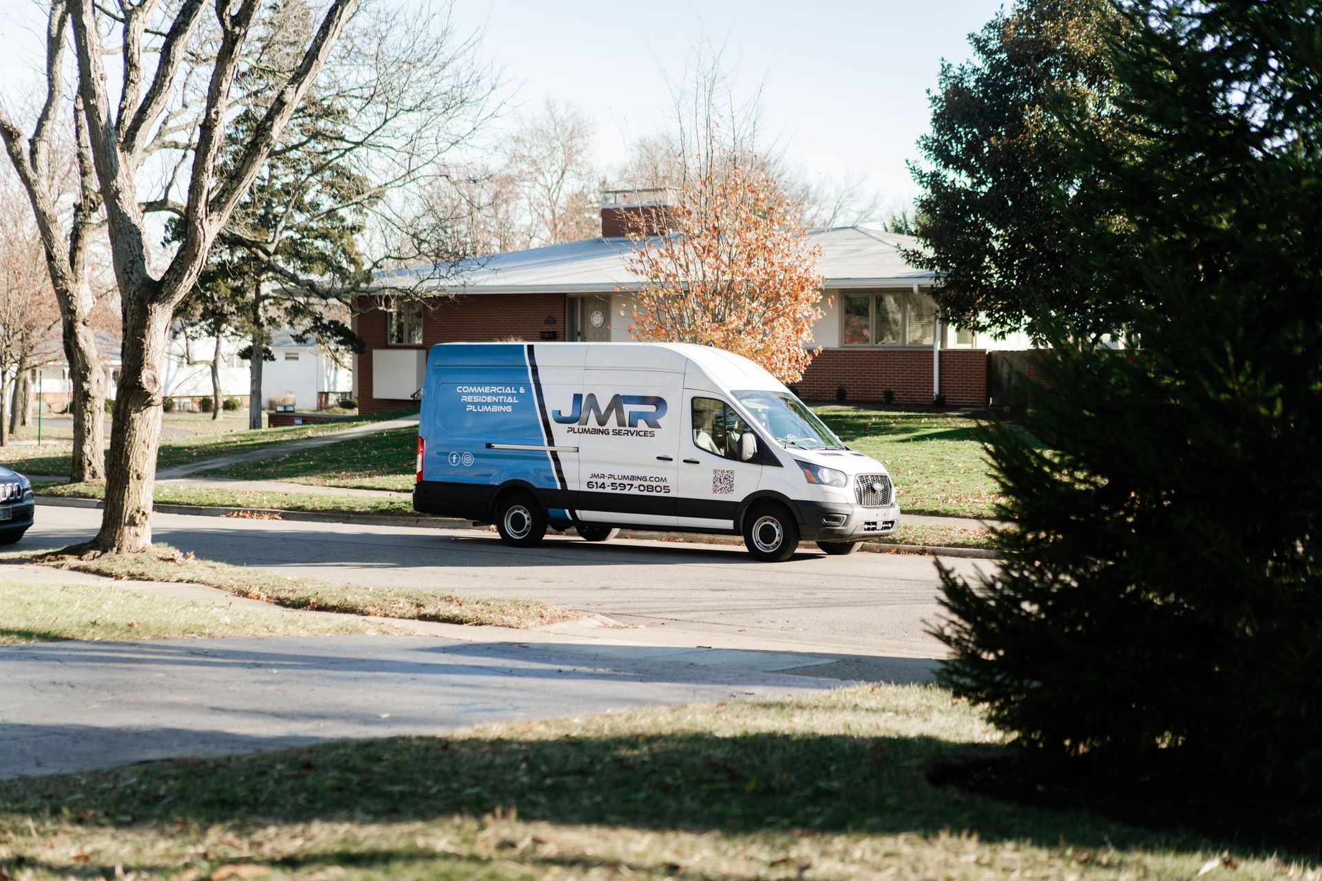 A white and blue JMR plumbing service van parked on a suburban street in front of a house.