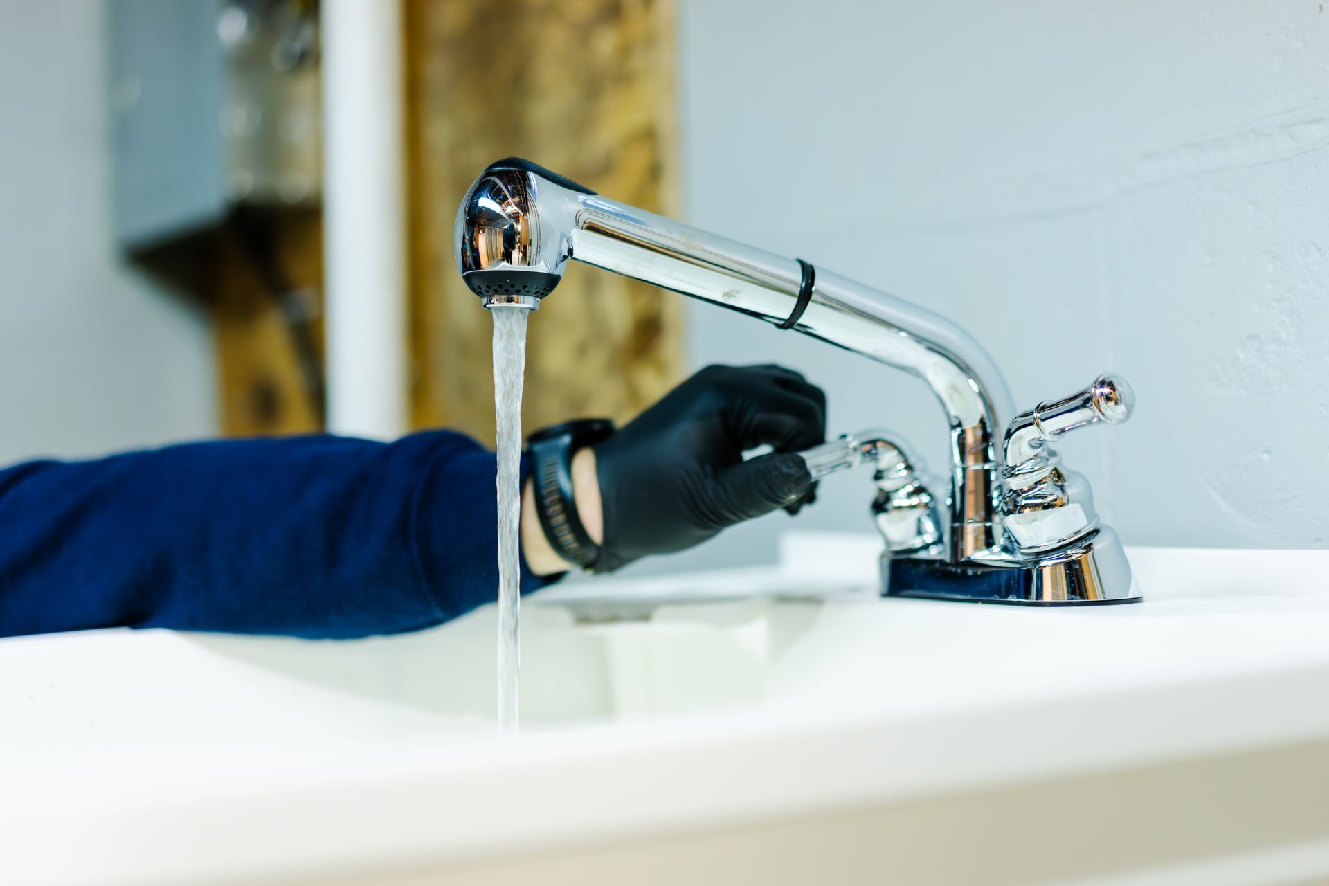A hand in a black glove adjusts the handle of a chrome kitchen faucet, with water running into a white sink.