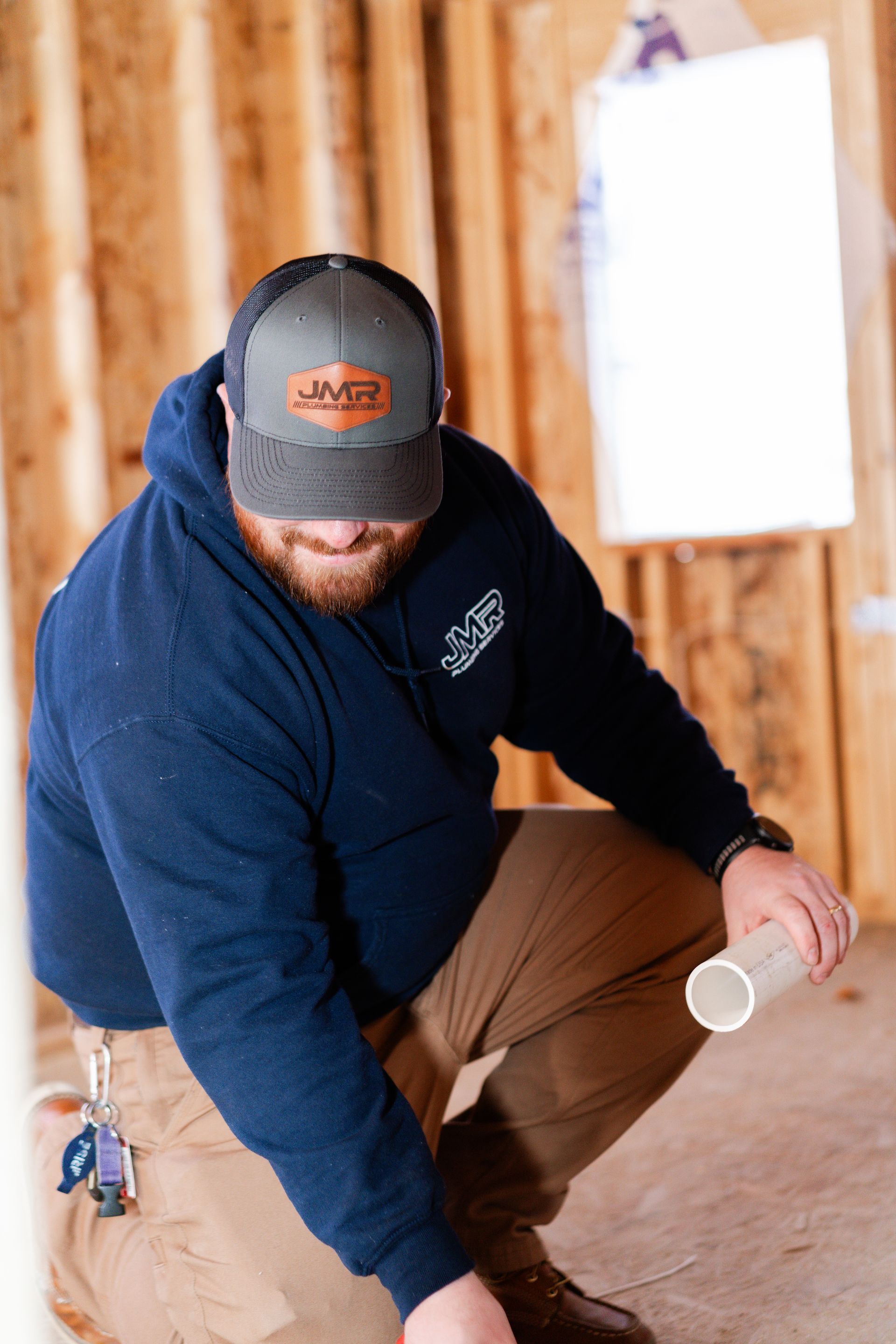 A person in a blue hoodie and gray baseball cap crouches in an unfinished building, holding a short section of pipe.