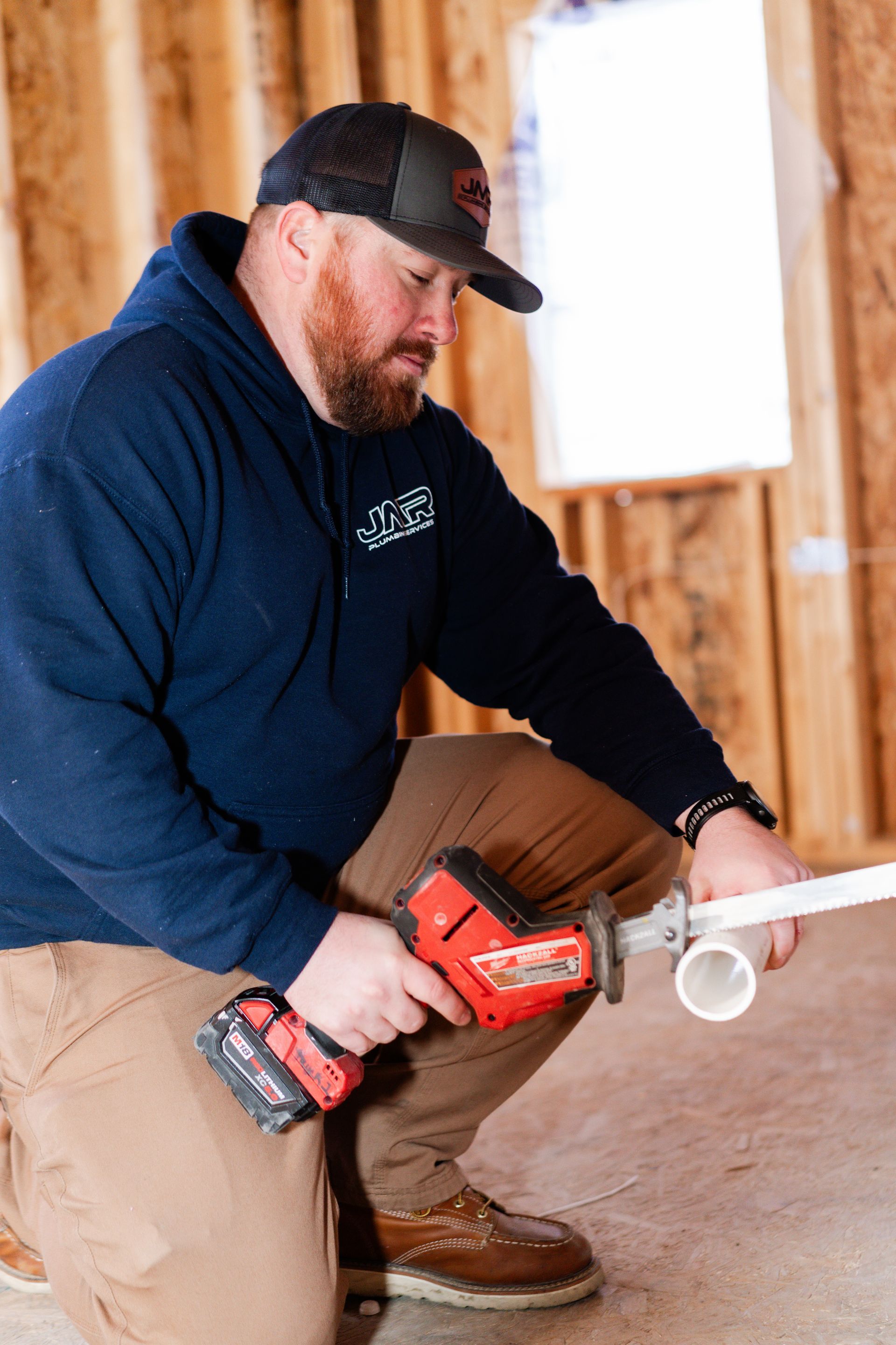 A person in a blue hoodie and cap kneels in a wooden framed building, using a cordless reciprocating saw to cut a pipe.