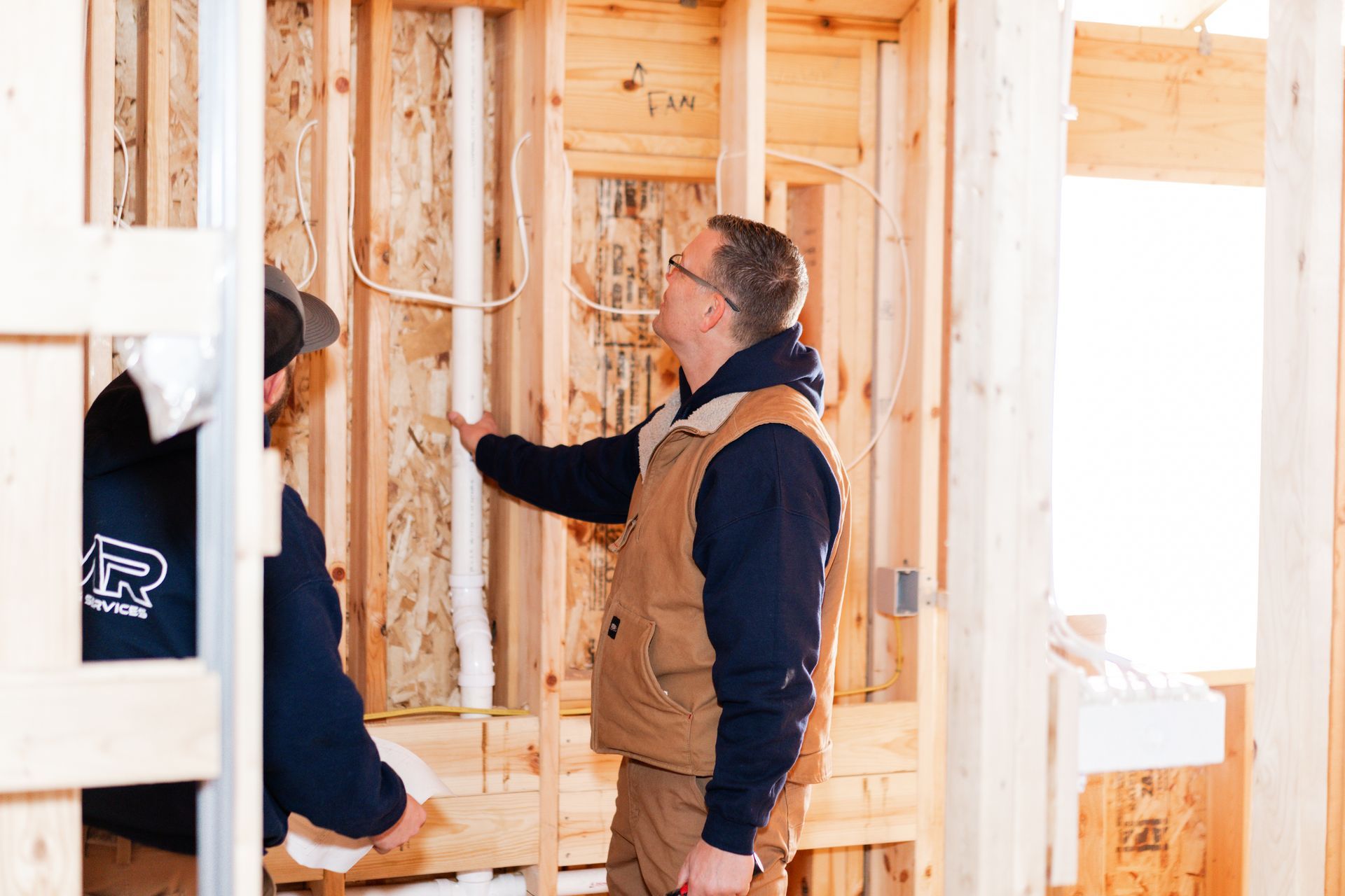 Two professionals in a construction site inspect a vertical white PVC pipe installed between wooden wall studs.