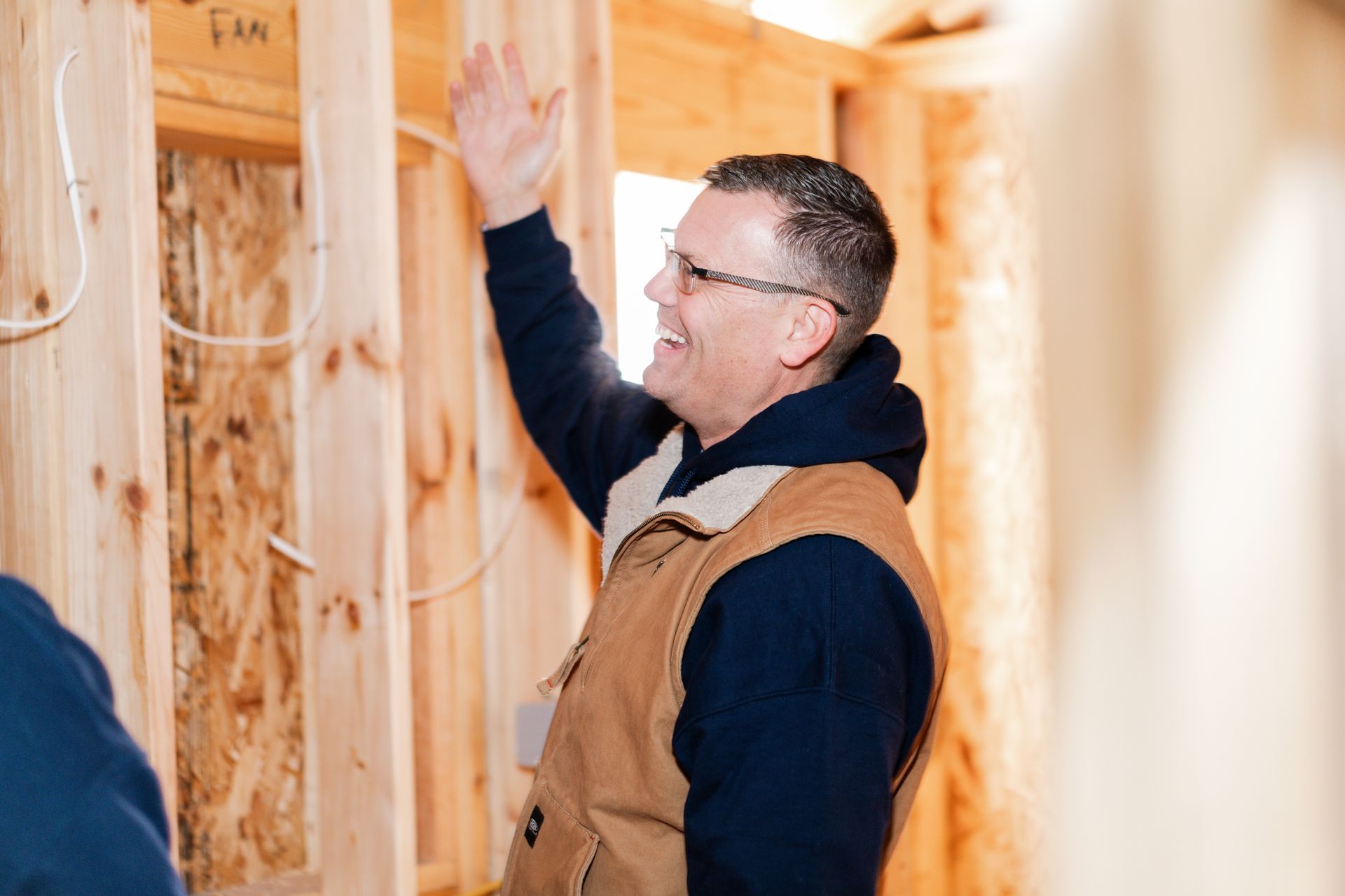 A person wearing glasses and a tan vest smiles and gestures toward wooden wall studs in a room under construction.