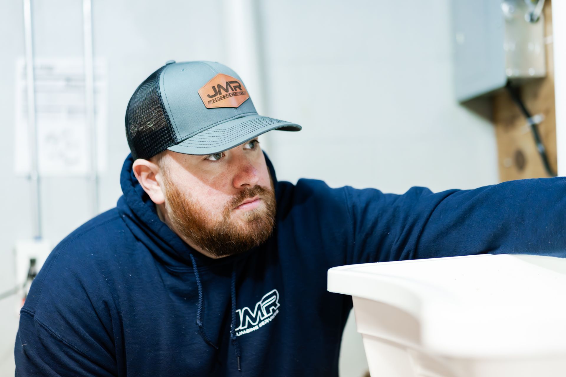 A professional in a navy hoodie and grey cap inspects plumbing equipment in an indoor setting.
