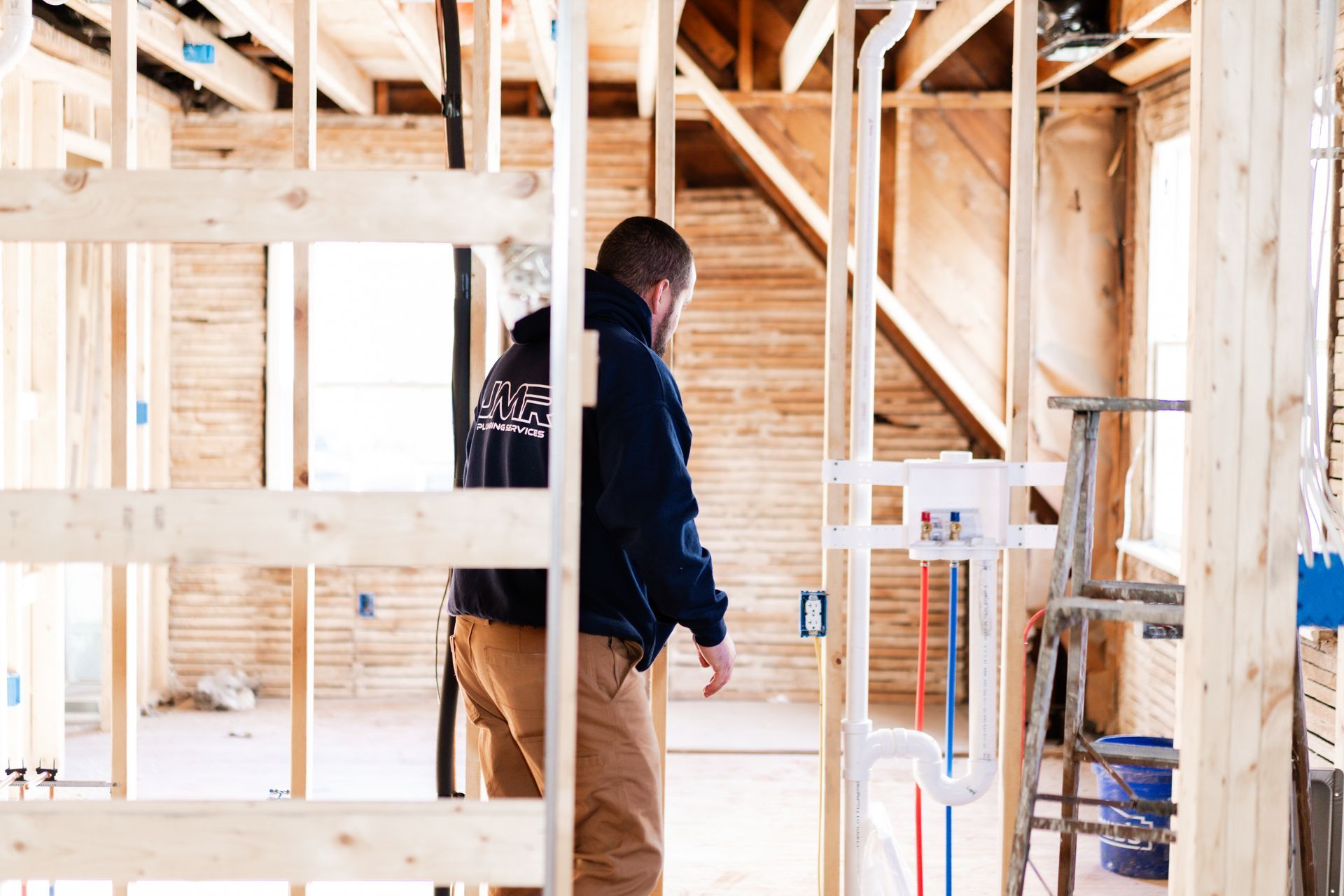 A person in a dark jacket and tan work pants inspects plumbing pipes inside the wooden frame of a construction site.