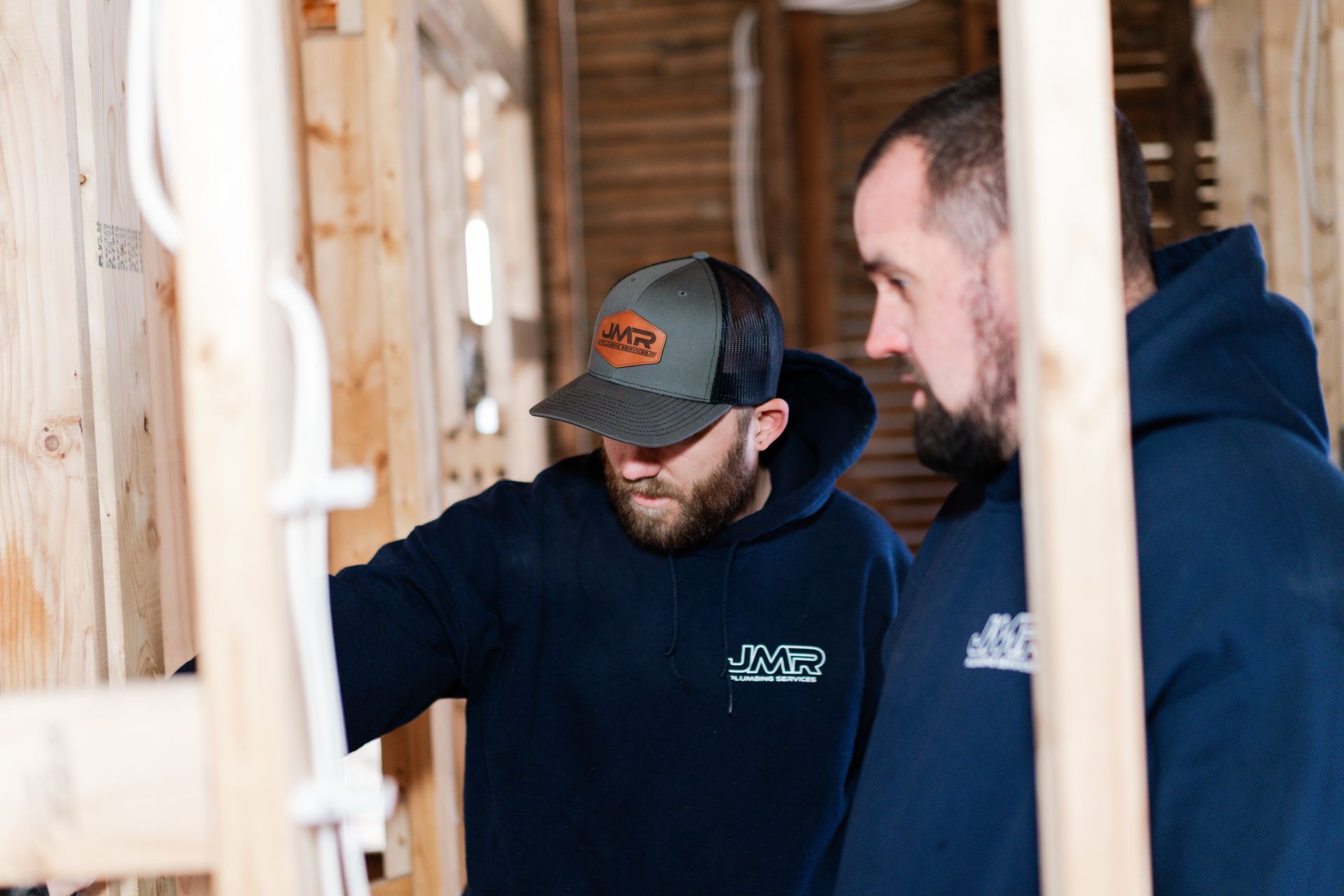 Two people in dark blue hoodies standing in an unfinished room with wooden wall studs, examining building work.