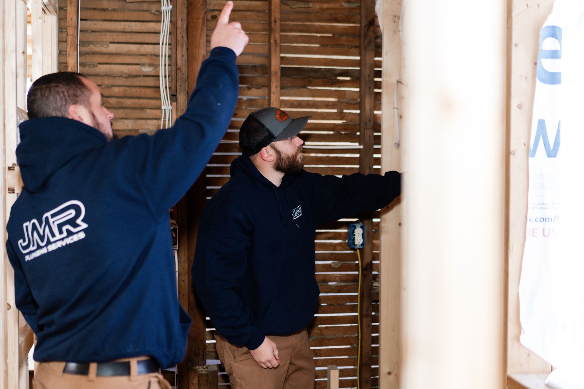 Two workers in navy hoodies examine a wall frame with wooden slats, one pointing toward the ceiling.