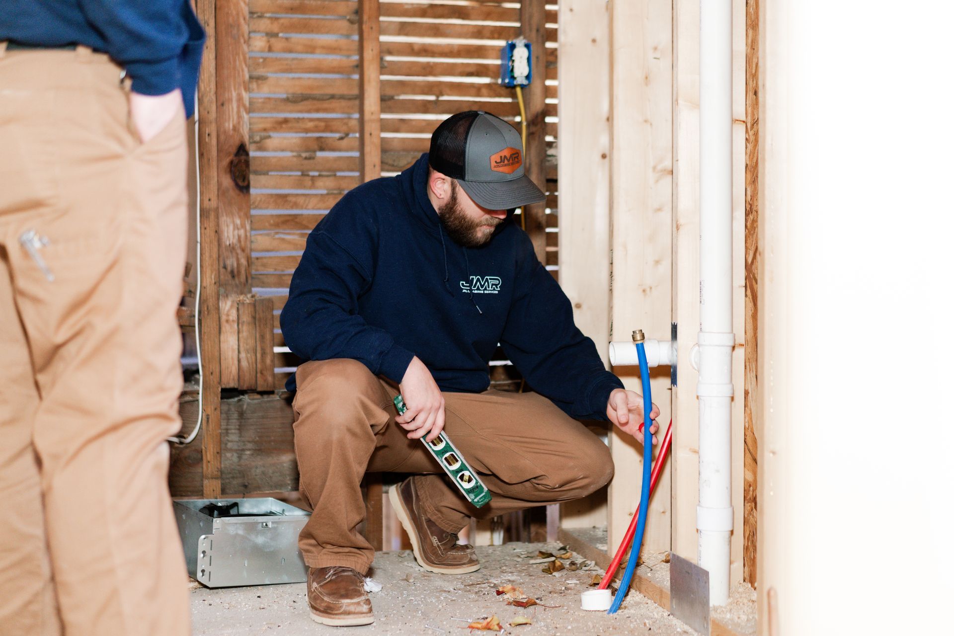 A person in a blue hoodie and tan work pants kneels to install red and blue plumbing pipes in an unfinished wall frame.