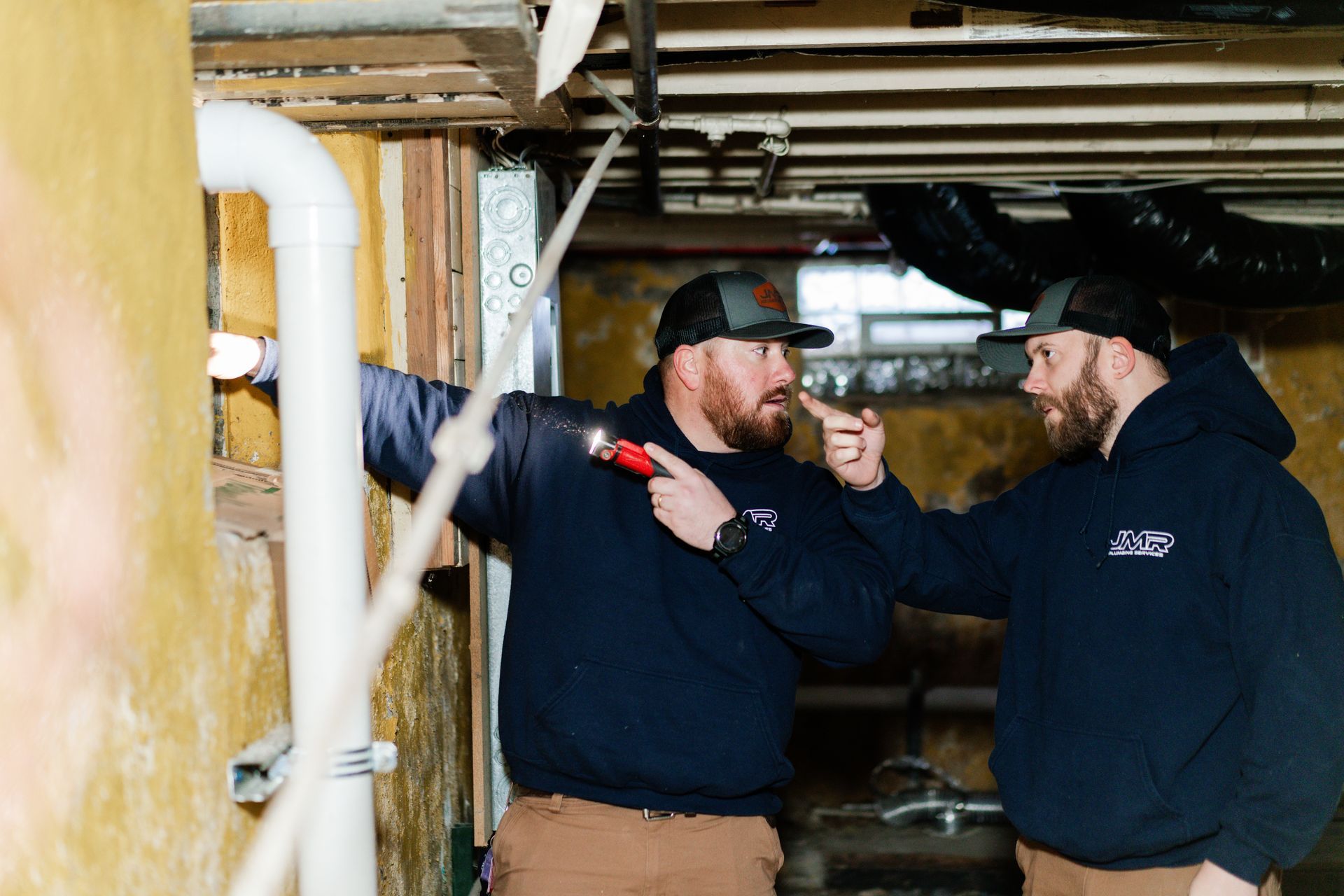 Two professionals in dark uniforms inspect a crawlspace, pointing at a white pipe and wall surface with a flashlight.