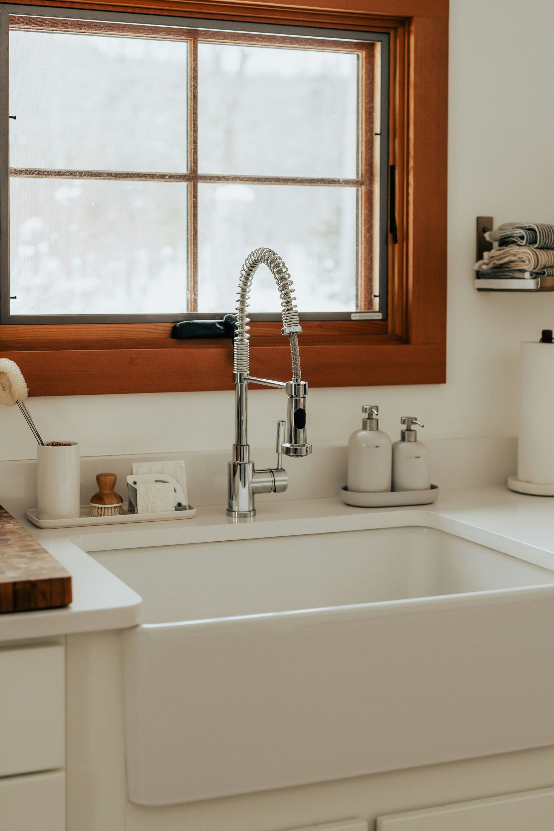 A white farmhouse kitchen sink with a chrome faucet below a window with a warm wood frame.
