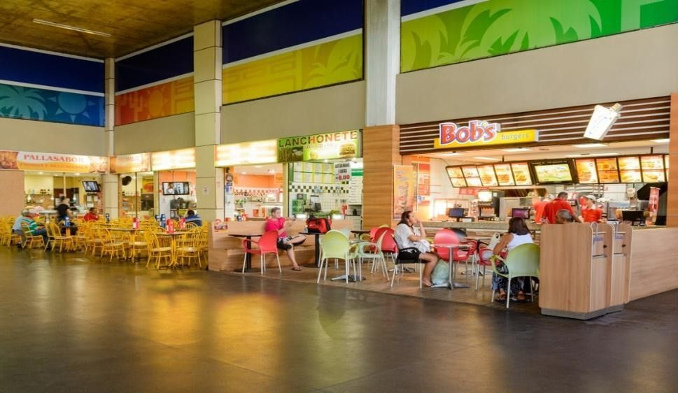 Praça de alimentação com diversos vendedores, mesas e pessoas comendo. Paredes coloridas, iluminação brilhante.