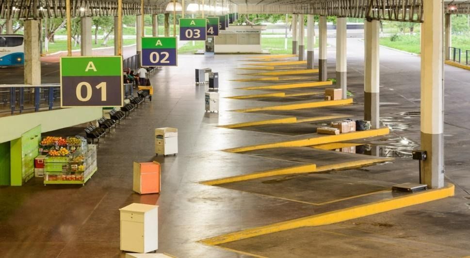 Terminal rodoviário vazio com plataformas numeradas demarcadas por linhas amarelas. Placas verdes e azuis suspensas.