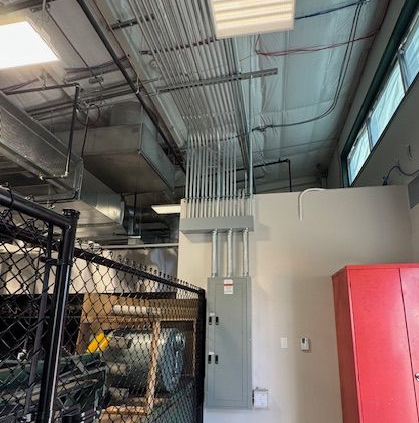 Electrical panel with conduits running to ceiling. Indoor setting with red cabinet and chain link fence.