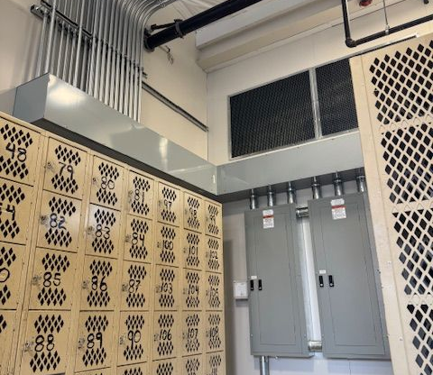 A room with lockers, electrical boxes, and conduits along the ceiling; numbers on the lockers.